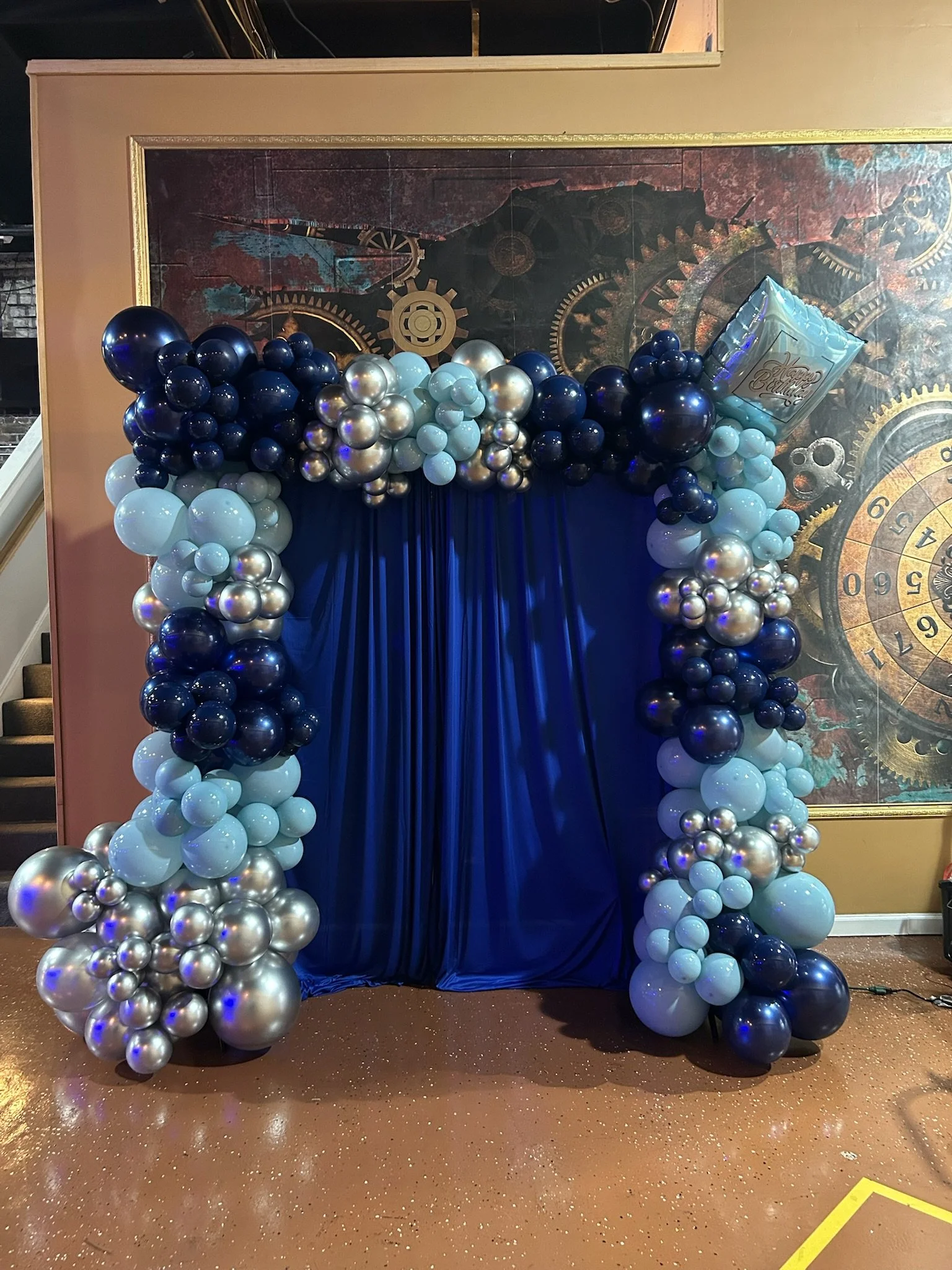 A decorative balloon arch with shades of blue, silver, and black balloons, set against a blue curtain backdrop and a steampunk-themed mural with gears and clock faces.