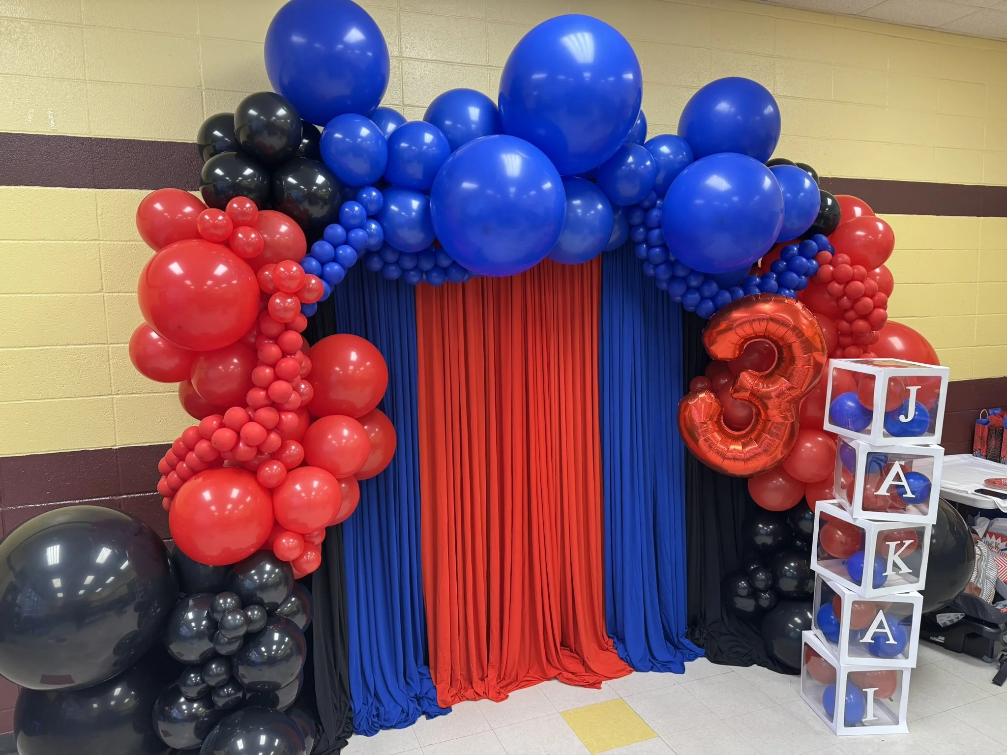 Colorful balloon decoration in red, black, and blue with a red curtain backdrop and a white cube display with letter blocks spelling out 'J', 'K', and 'A'.