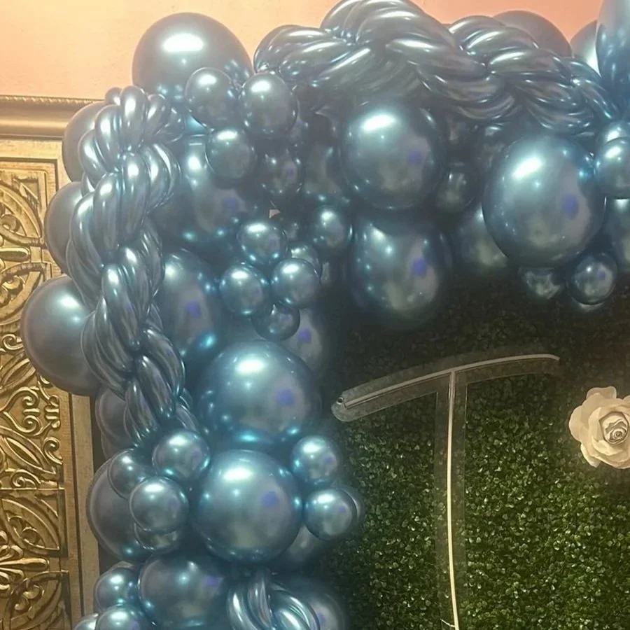 Silver balloons arranged in an arch with a green leafy backdrop and a large metallic number '1' in the background, with a white rose to the right.