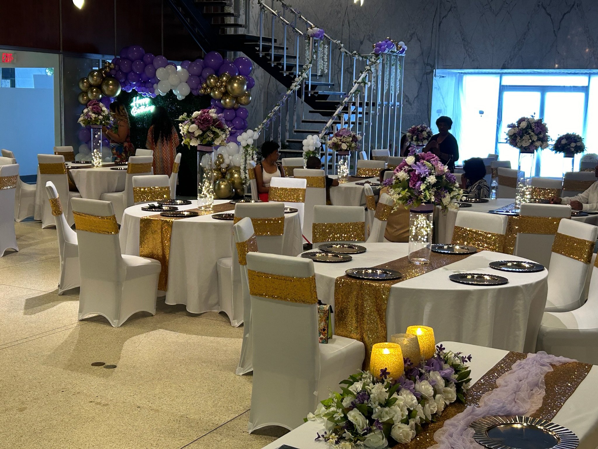 Wedding reception area with round tables covered in white tablecloths, decorated with gold glitter runners, and adorned with large floral arrangements of purple and white flowers. Purple, white, and gold balloons form an arch over a small backdrop with the illuminated word 'Happy.' Candles and string lights decorate the tables. The room has a marble wall, a staircase, and large windows with white curtains.