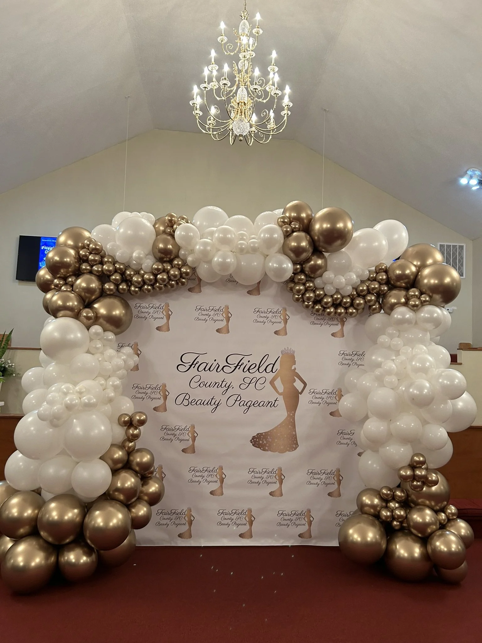 Balloon arch with white and gold balloons in front of a backdrop that says 'FairField County, SC Beauty Pageant' along with a silhouette of a woman in a crown.