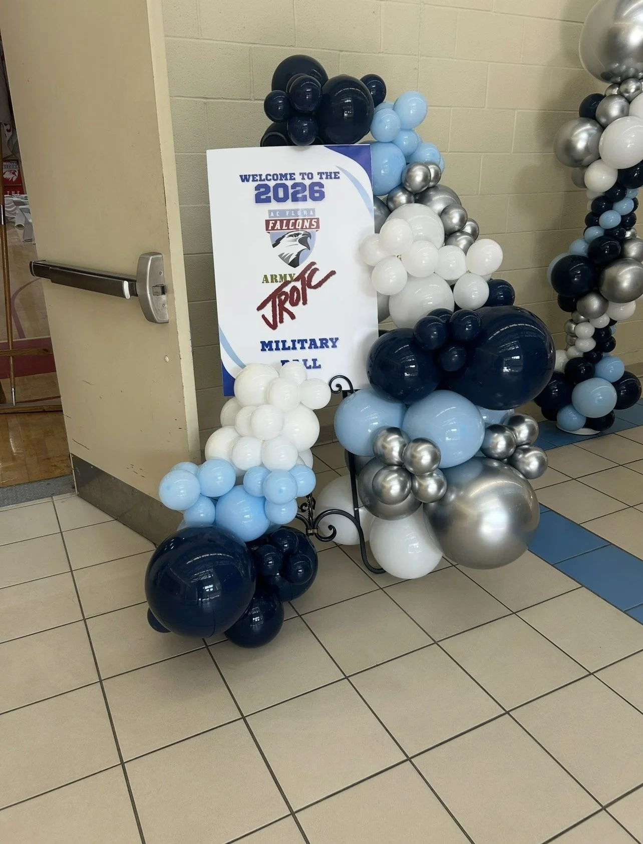 Balloon arrangement with white, blue, navy, and silver balloons near a sign that reads 'Welcome to the 2026 AC Flora Falcons Army JROTC Military Ball' in a school or community center hallway.