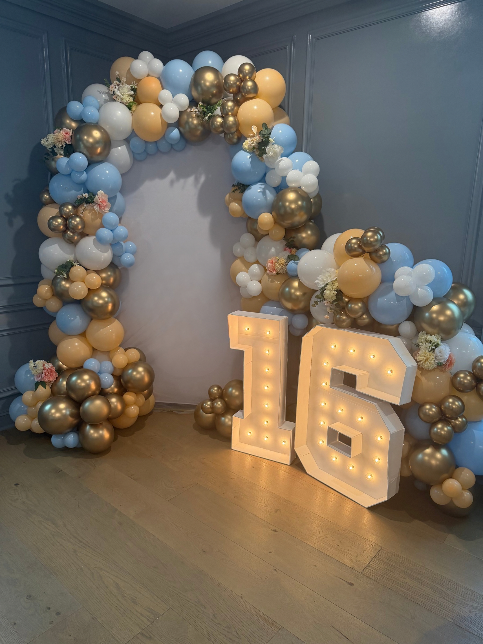 Decorative balloon arch with gold, white, blue, and beige balloons, floral accents, and illuminated '16' marquee on a wooden floor with gray wall background.
