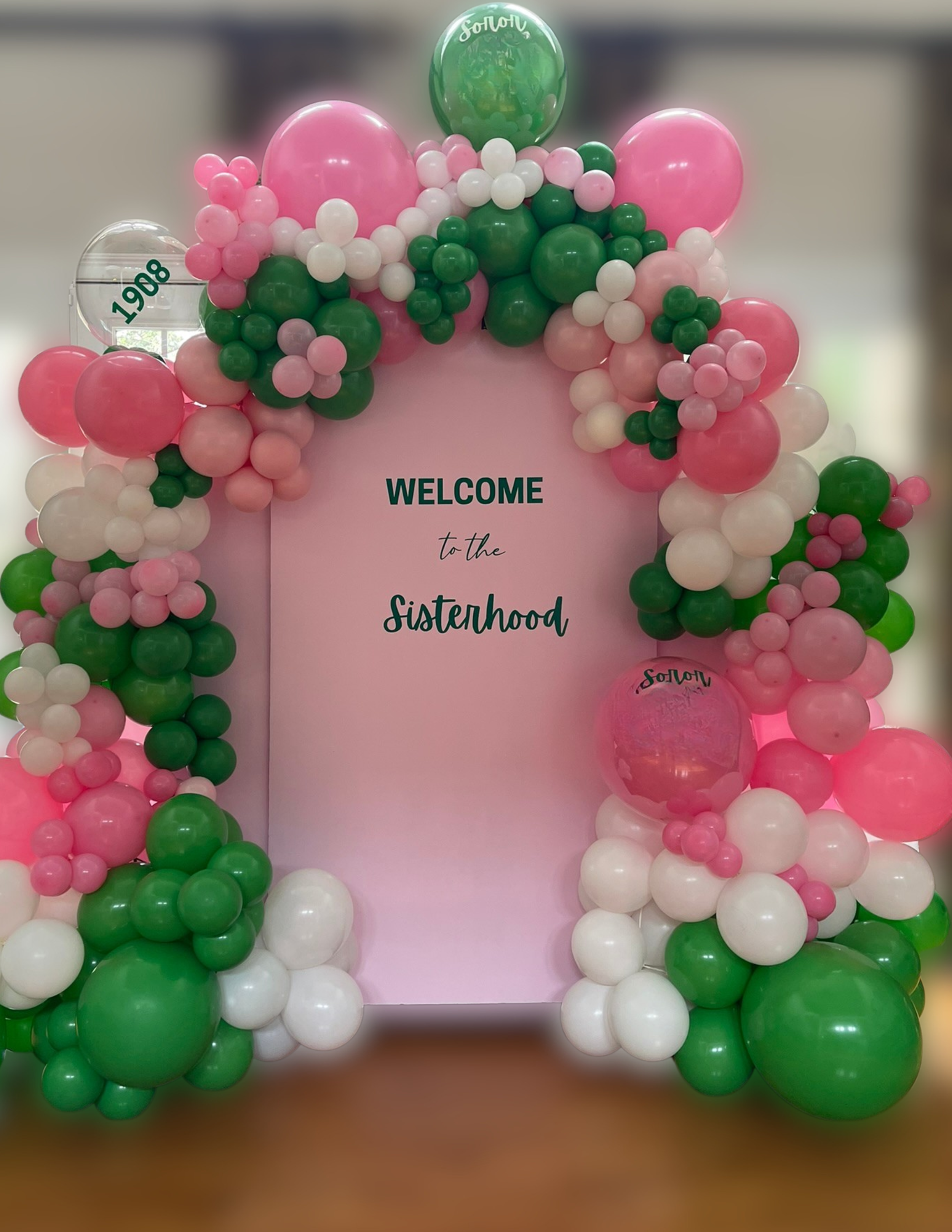 Colorful balloon arch with pink, green, white, and clear balloons surrounding a pink backdrop that reads "Welcome to the Sisterhood."