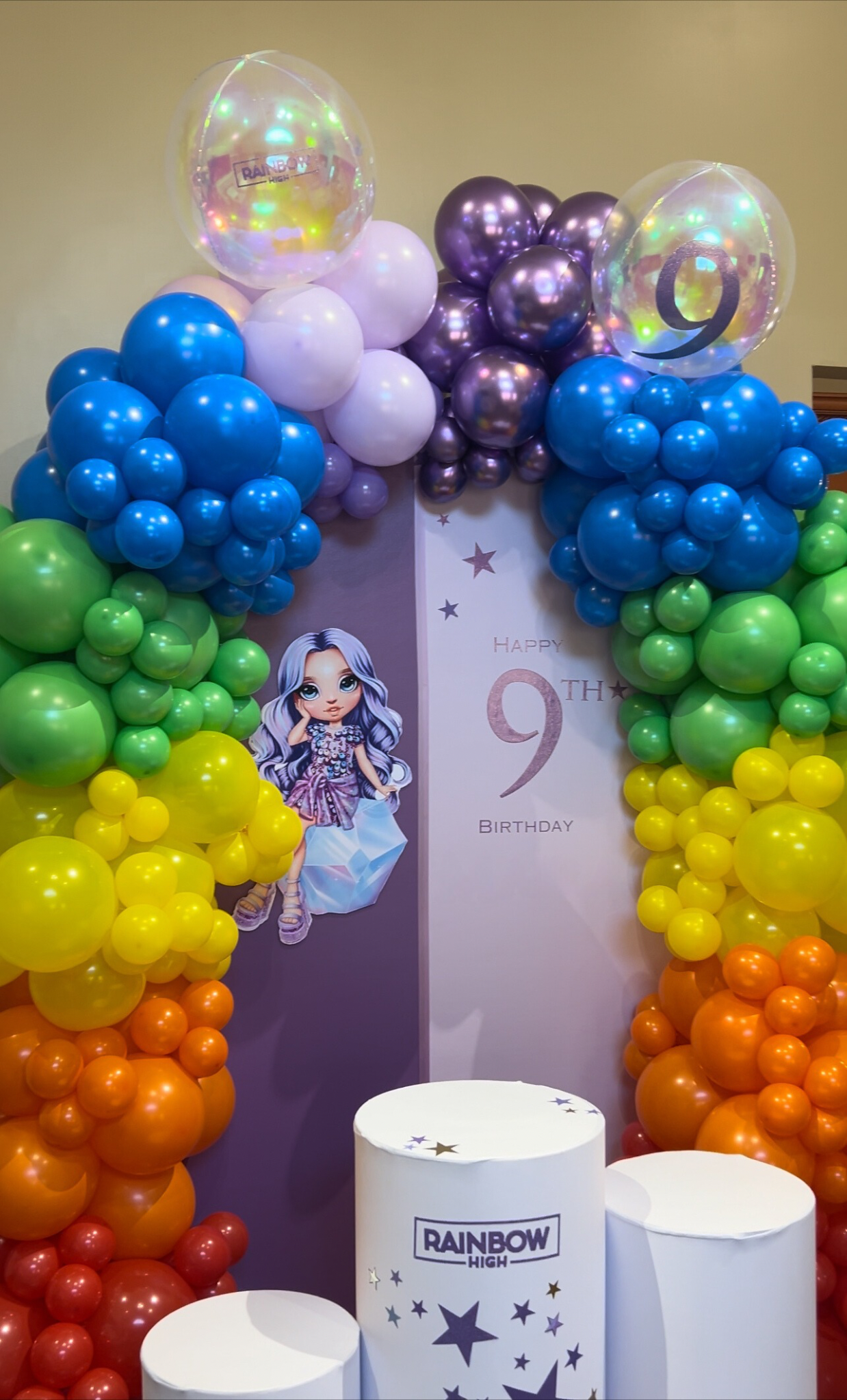 Colorful balloon arch with purple, blue, green, yellow, orange, and red balloons, a birthday backdrop with a cartoon girl illustration, and white cylindrical platforms with star designs and the words "Rainbow High" for a 9th birthday celebration.