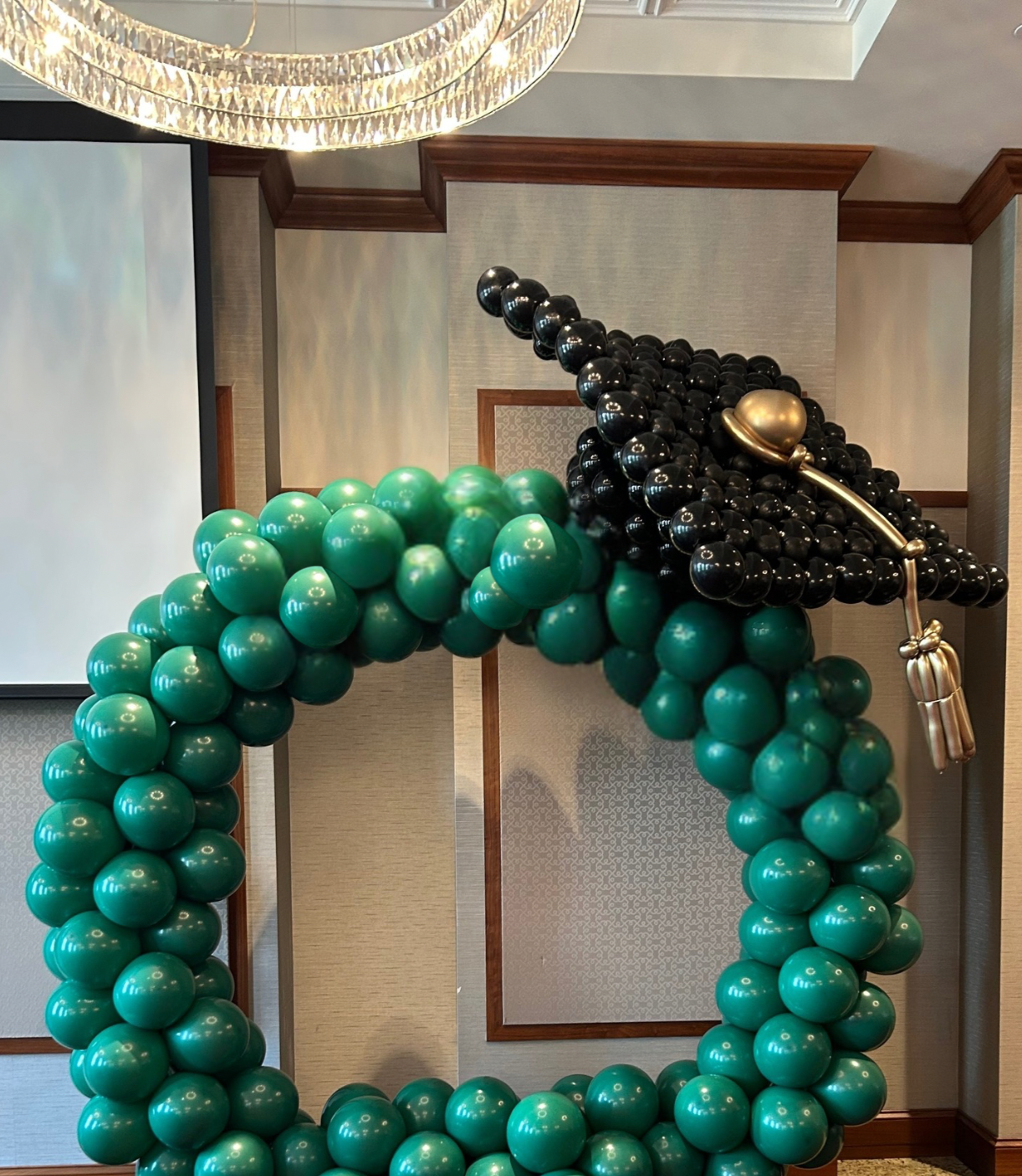 Balloon sculpture of a grand piano, made with green and black balloons, decorated with a gold speaker and a gold microphone, situated in a room with beige walls and a chandelier.