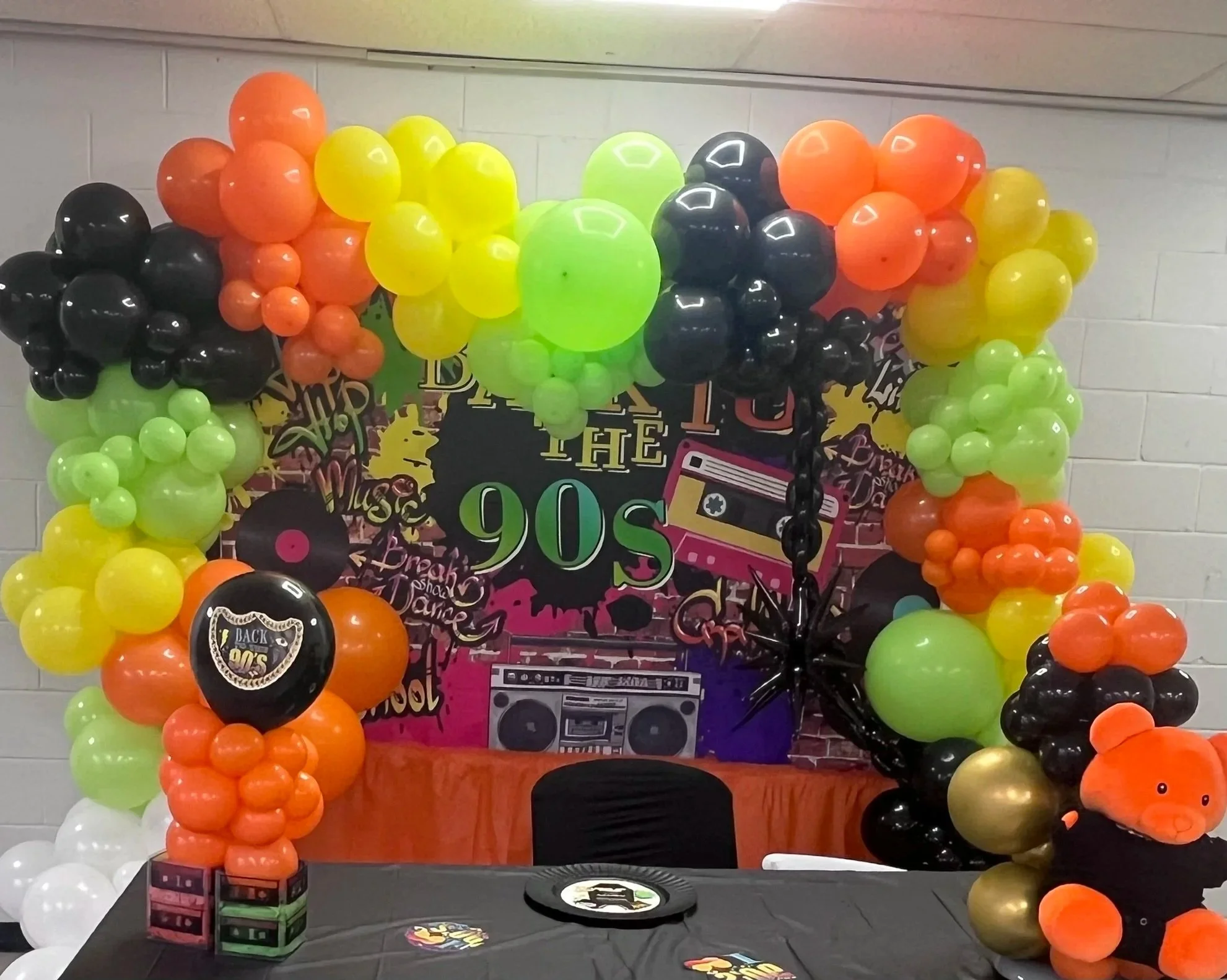 Colorful balloon arrangement with black, yellow, orange, and white balloons framing a 1990s-themed party display. The display features a sign with the year '1990s,' a boombox, vinyl records, and a cassette tape, celebrating a 90s retro theme.