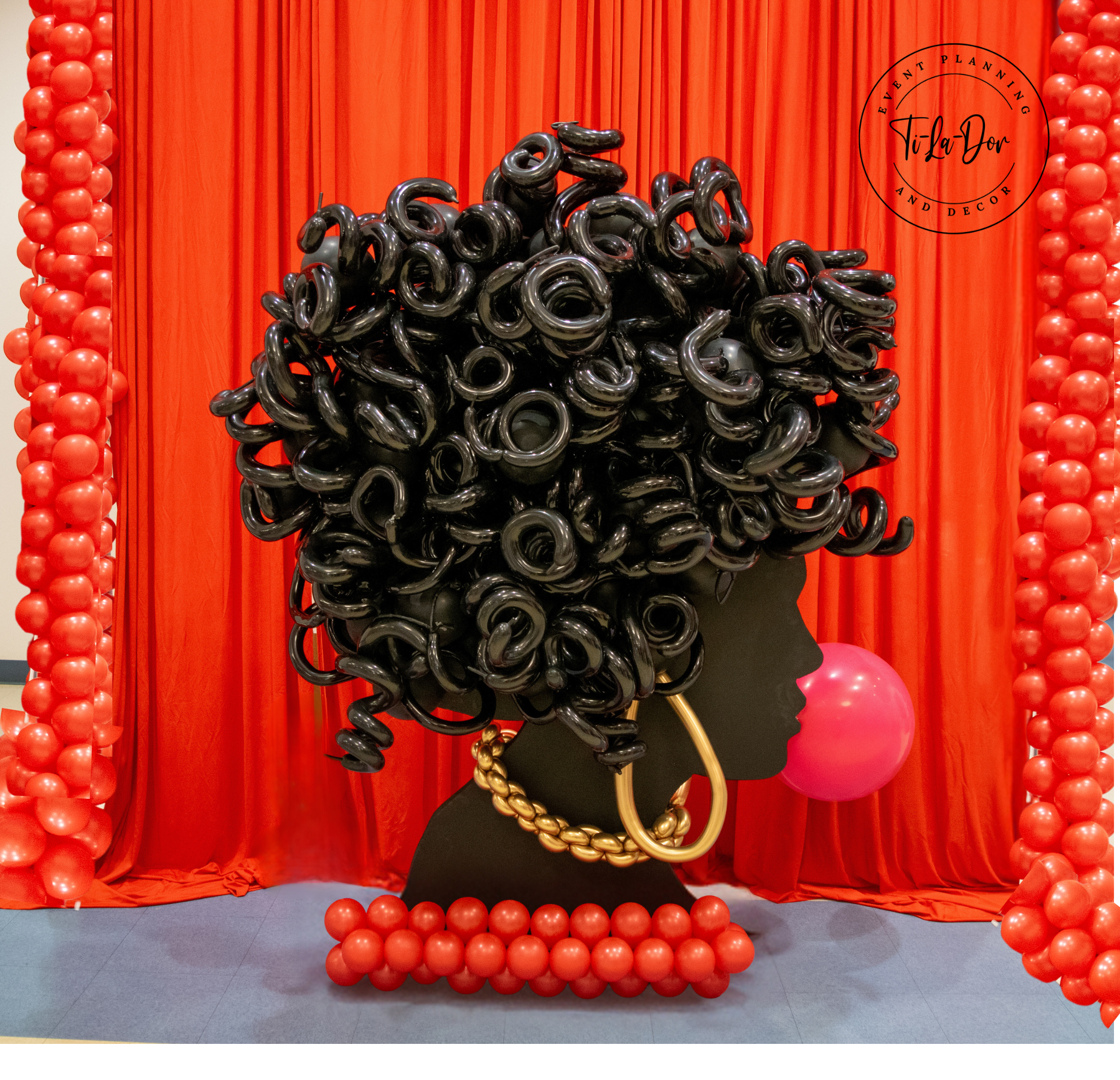 Decorative display with red balloons, a black mannequin head, and a large black and gold chain necklace, set against red curtains with a logo for event planning and decor.