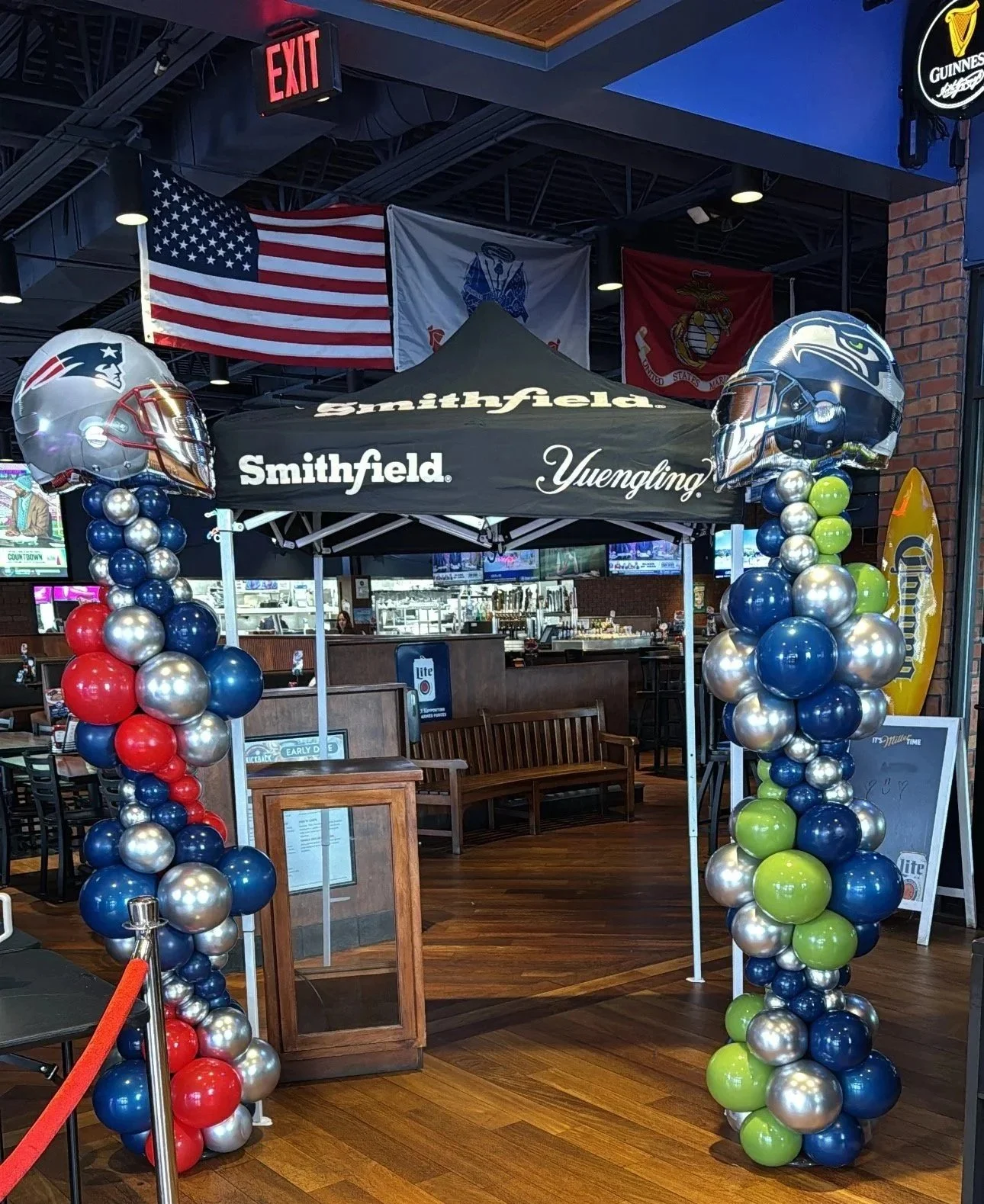 Entrance to a sports bar decorated with balloons in red, blue, green, and silver, featuring football helmets as balloon tops. Inside the bar, American flag and several military flags are displayed. A black tent with the Smithfield Yuengling logo marks the entrance.
