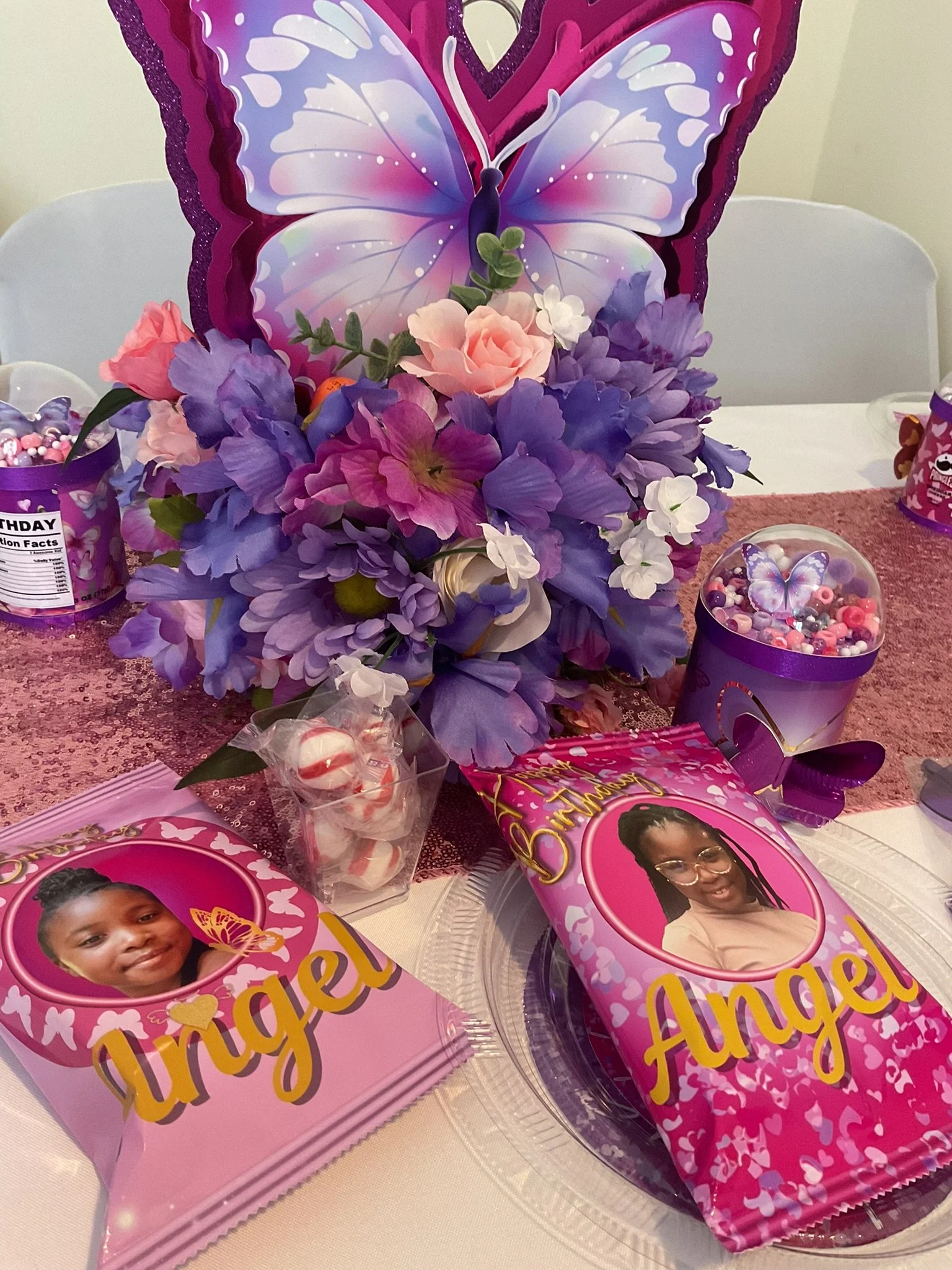 A birthday celebration setup with purple and pink theme, featuring a large butterfly decoration, floral arrangements, and party favors such as plates, cups, and goodie bags with images of a young girl's face and birthday messages.