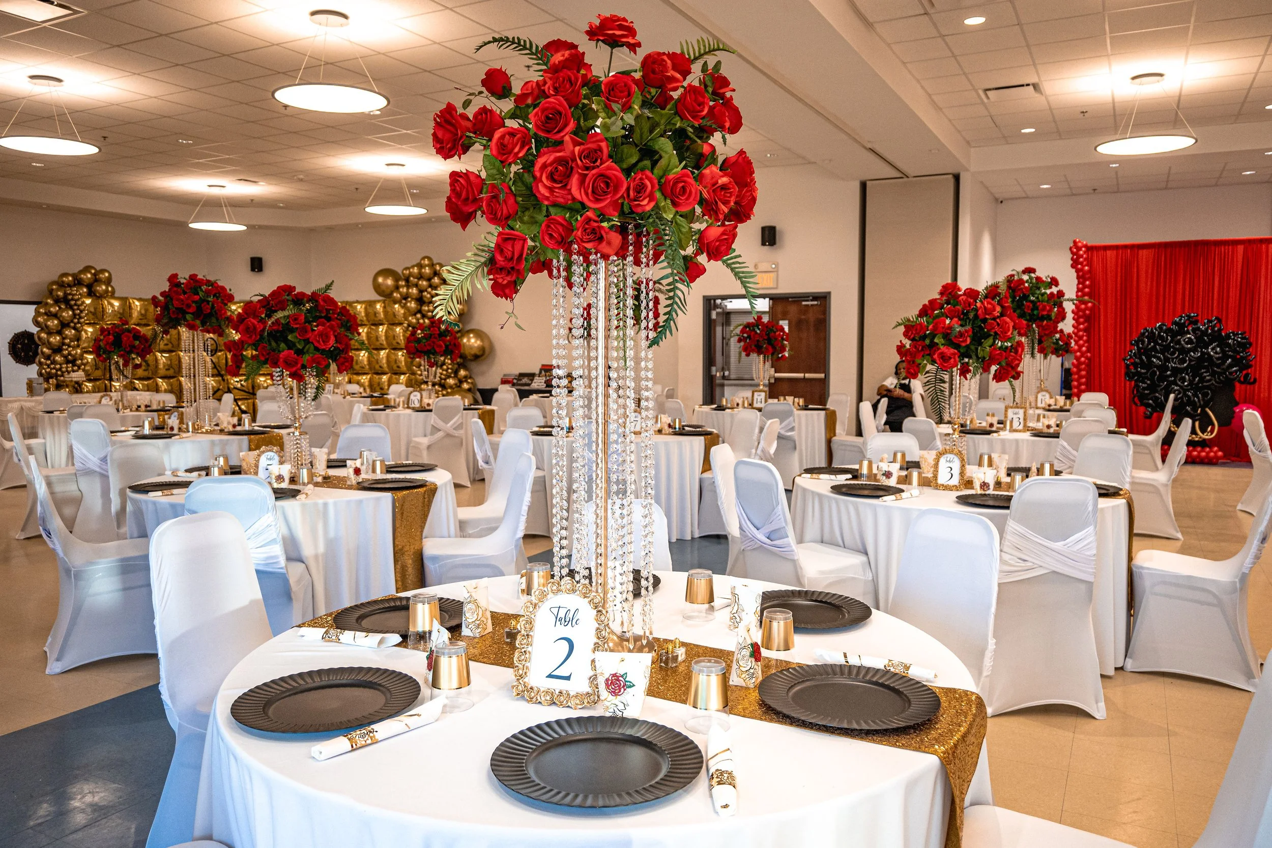 Indoor banquet hall decorated with large red rose floral arrangements and gold accents, round tables with black plates and gold and white table decorations, gold balloon wall backdrop, and a red curtain with balloon border.