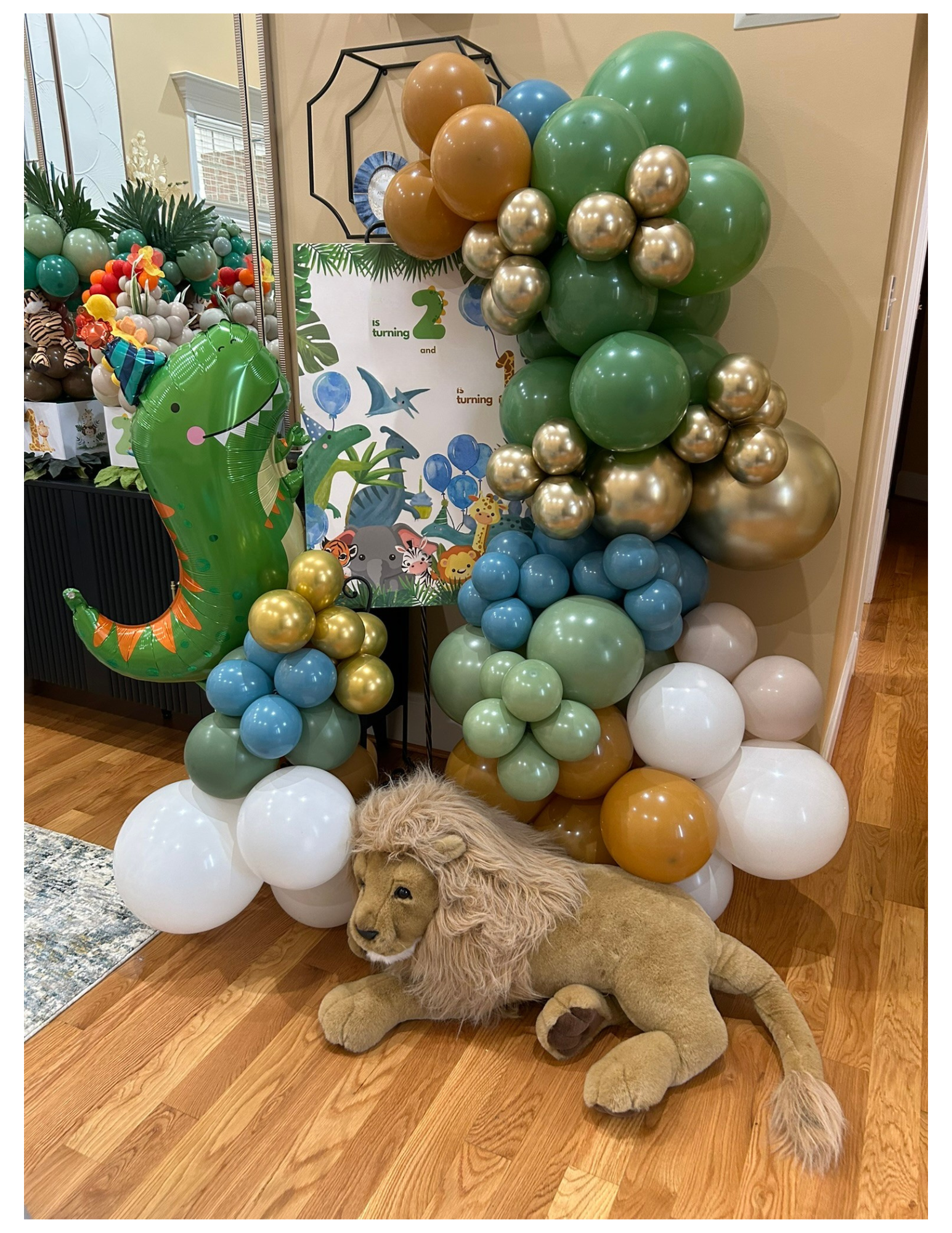 Party decoration with green, gold, blue, and white balloons arranged in an arch and cluster, featuring a jungle safari theme with a plush lion in front, toy animals, and a jungle-themed backdrop with animals and greenery.