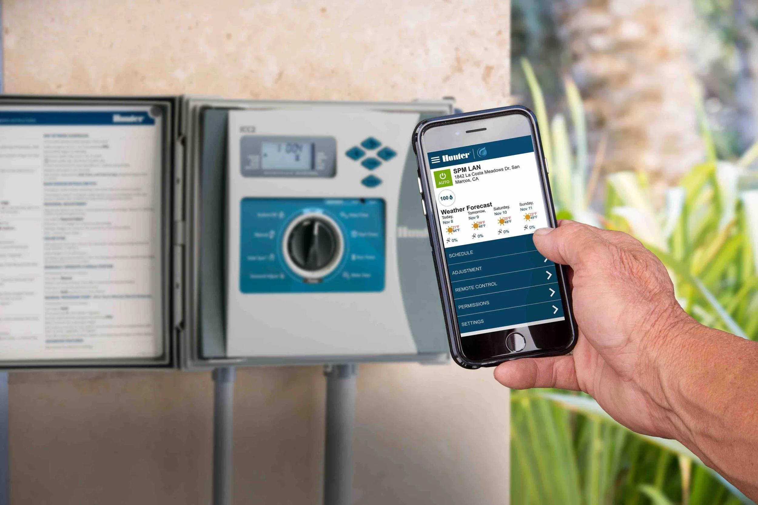 A man's hand holds a smartphone in fromt of an irrigation control box mounted on a stucco wall.