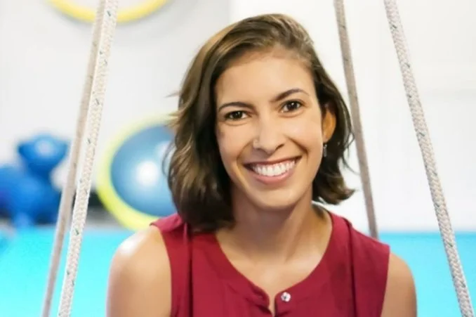 A smiling woman with shoulder-length brown hair, wearing a red sleeveless top, sitting on a swing in a fitness gym with weights and exercise balls in the background.