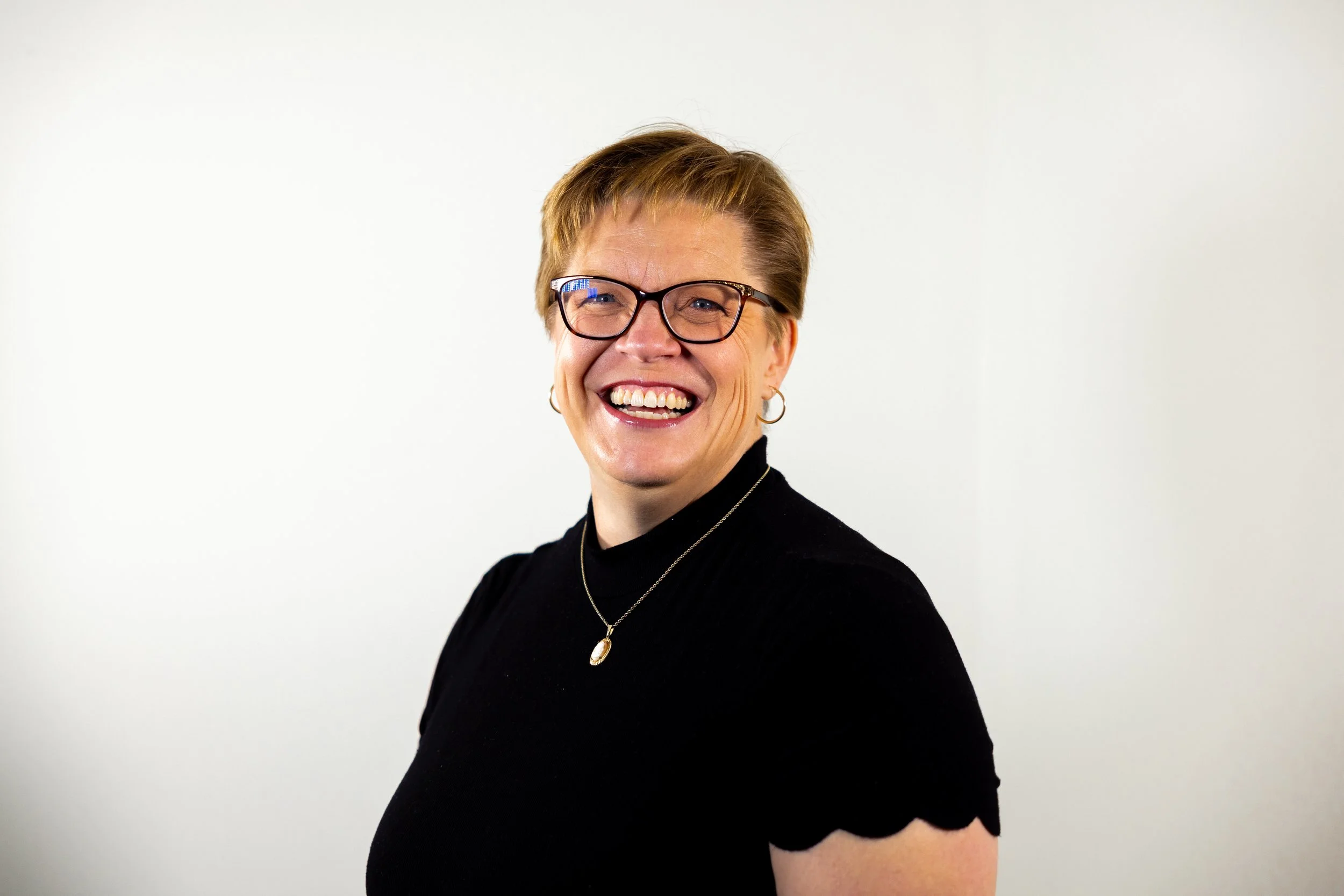 A smiling woman with short red hair, glasses, hoop earrings, and a black top, standing against a plain white background.