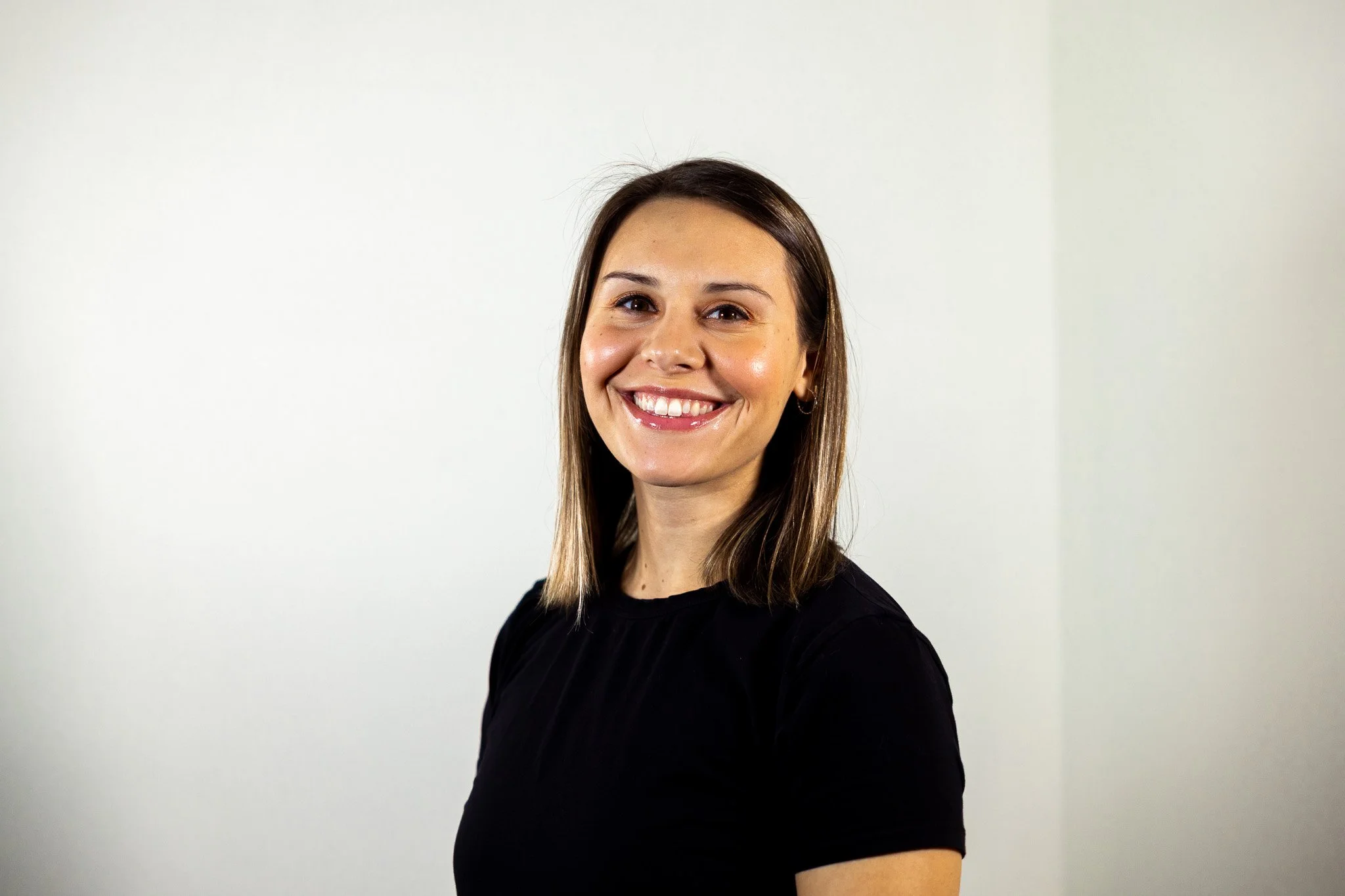 A smiling woman with shoulder-length brown hair and hoop earrings wearing a black shirt standing against a plain white background.