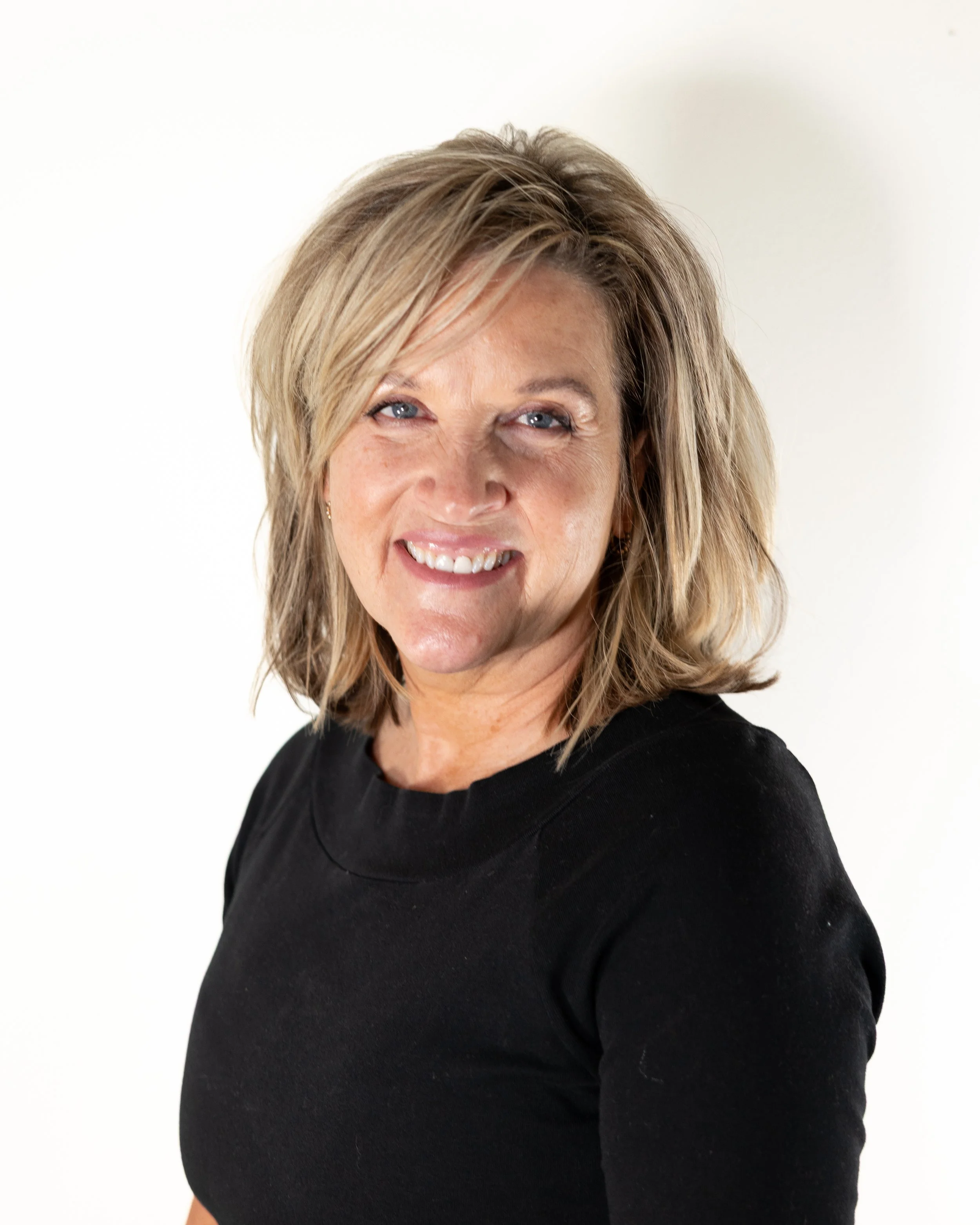 A woman with wavy blonde hair smiling in front of a plain white background.