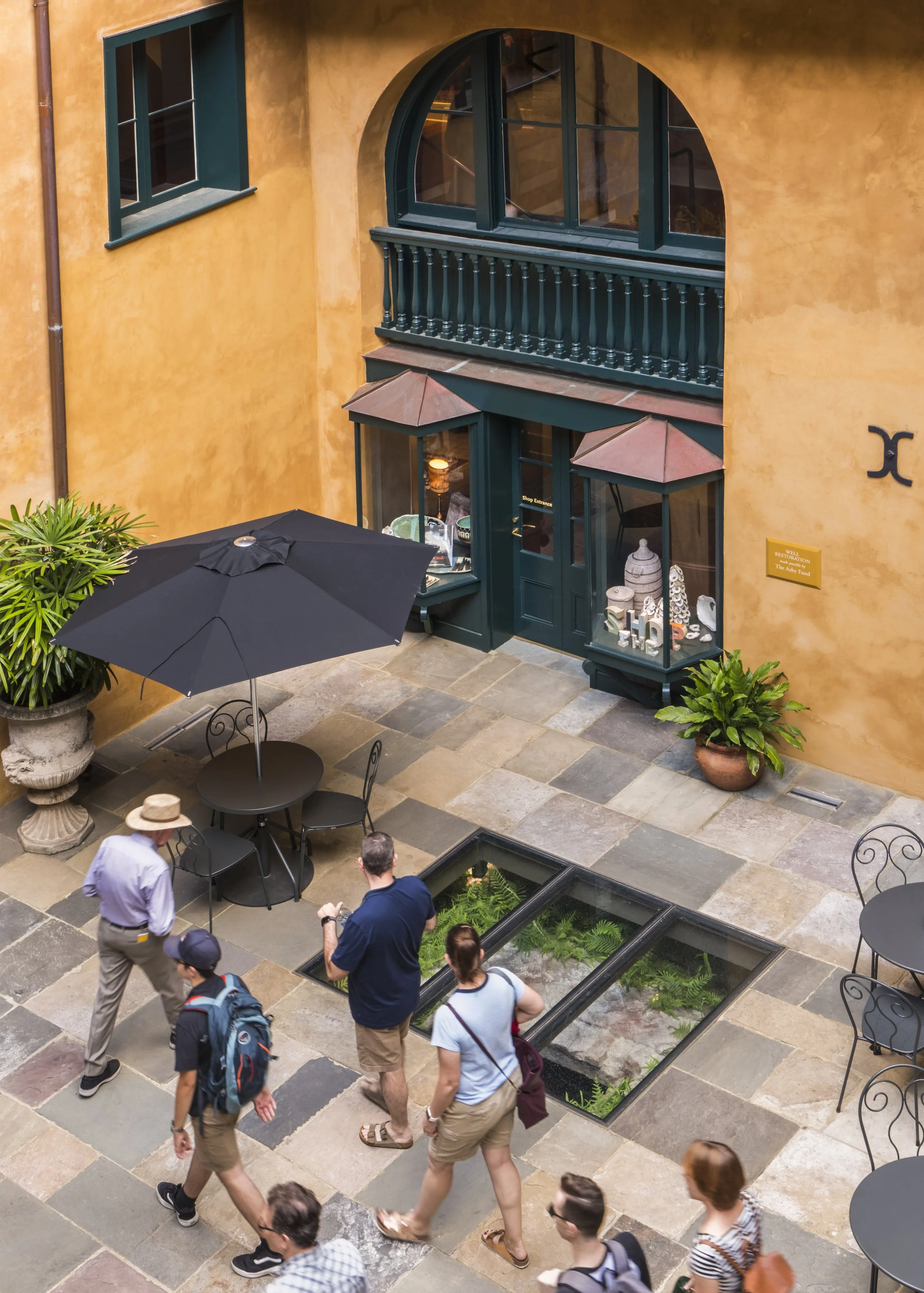 People walking through a courtyard with potted plants, a black umbrella with seating, and a shop with large windows on an orange wall.