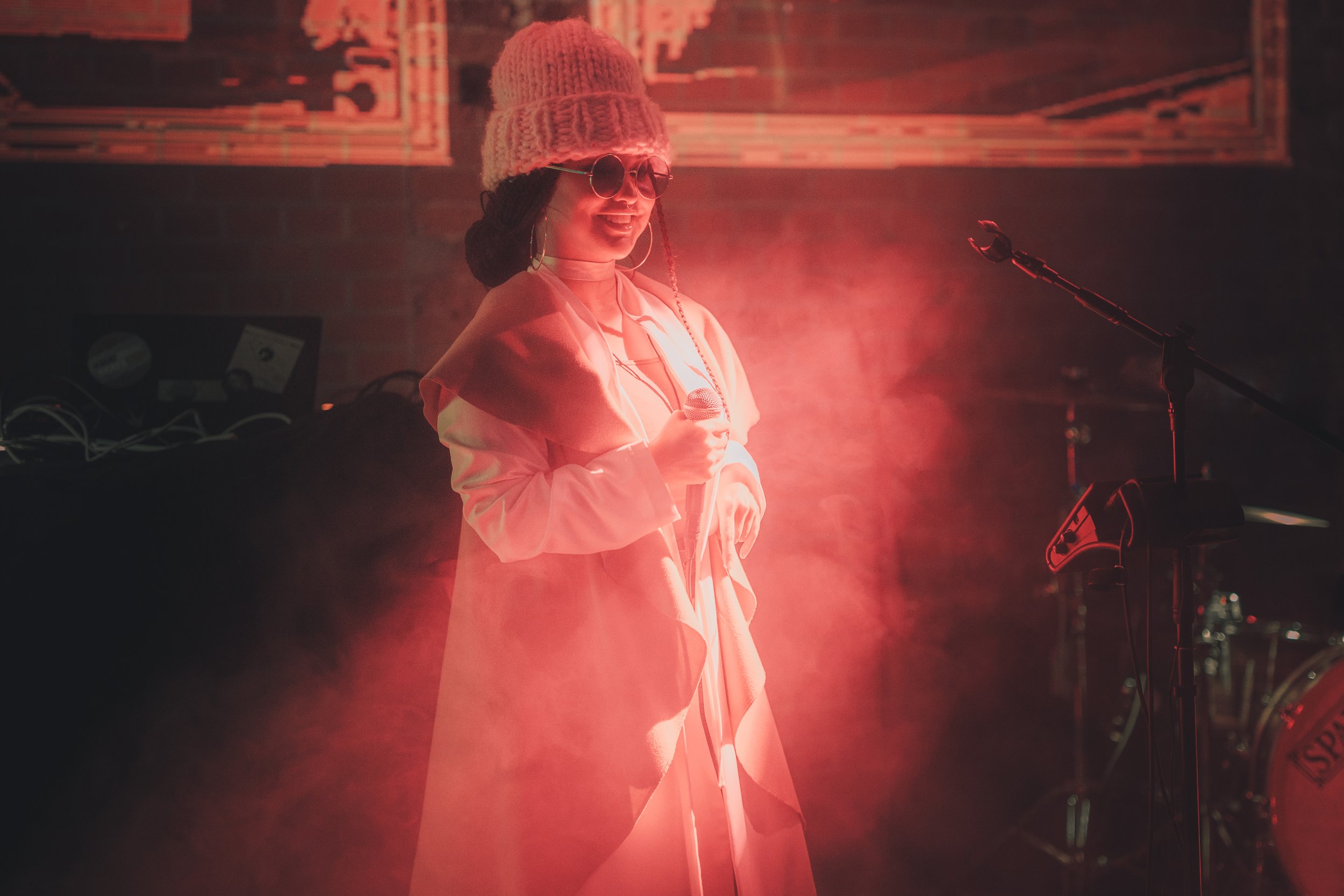 female singer on stage with a lot of fog holding microphone, waring all white and sunglasses partly lit by a red lightbeam