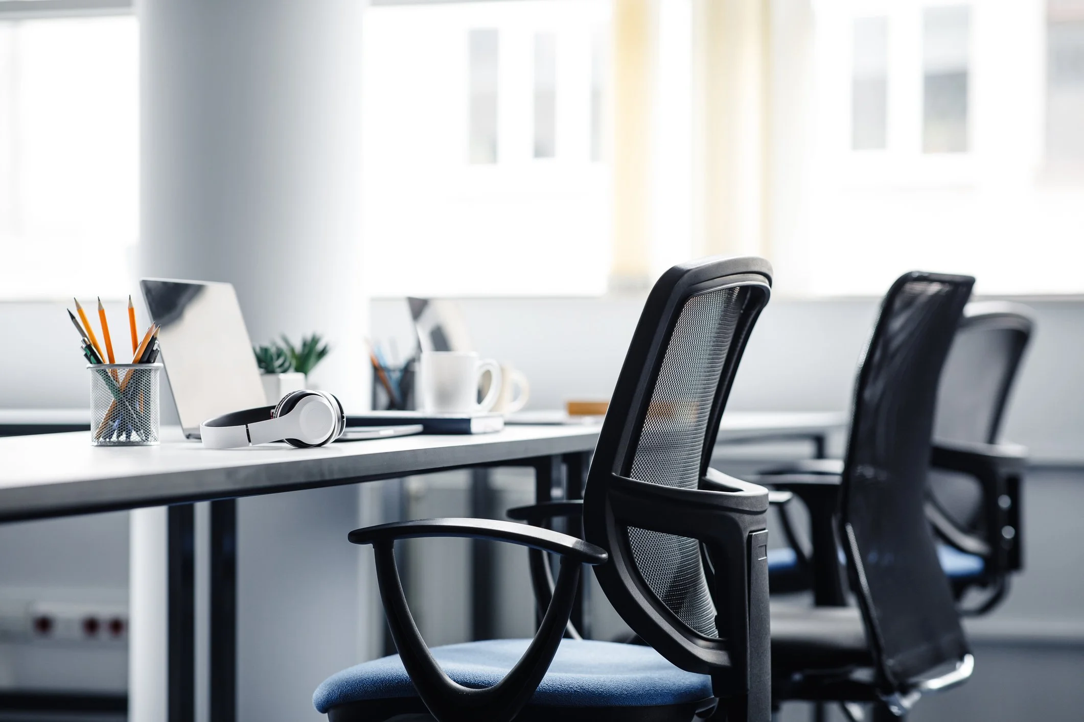 Office conference room with black mesh chairs around a white table, a container of pencils, a potted plant, a laptop, a white headset on the table, and large windows in the background.