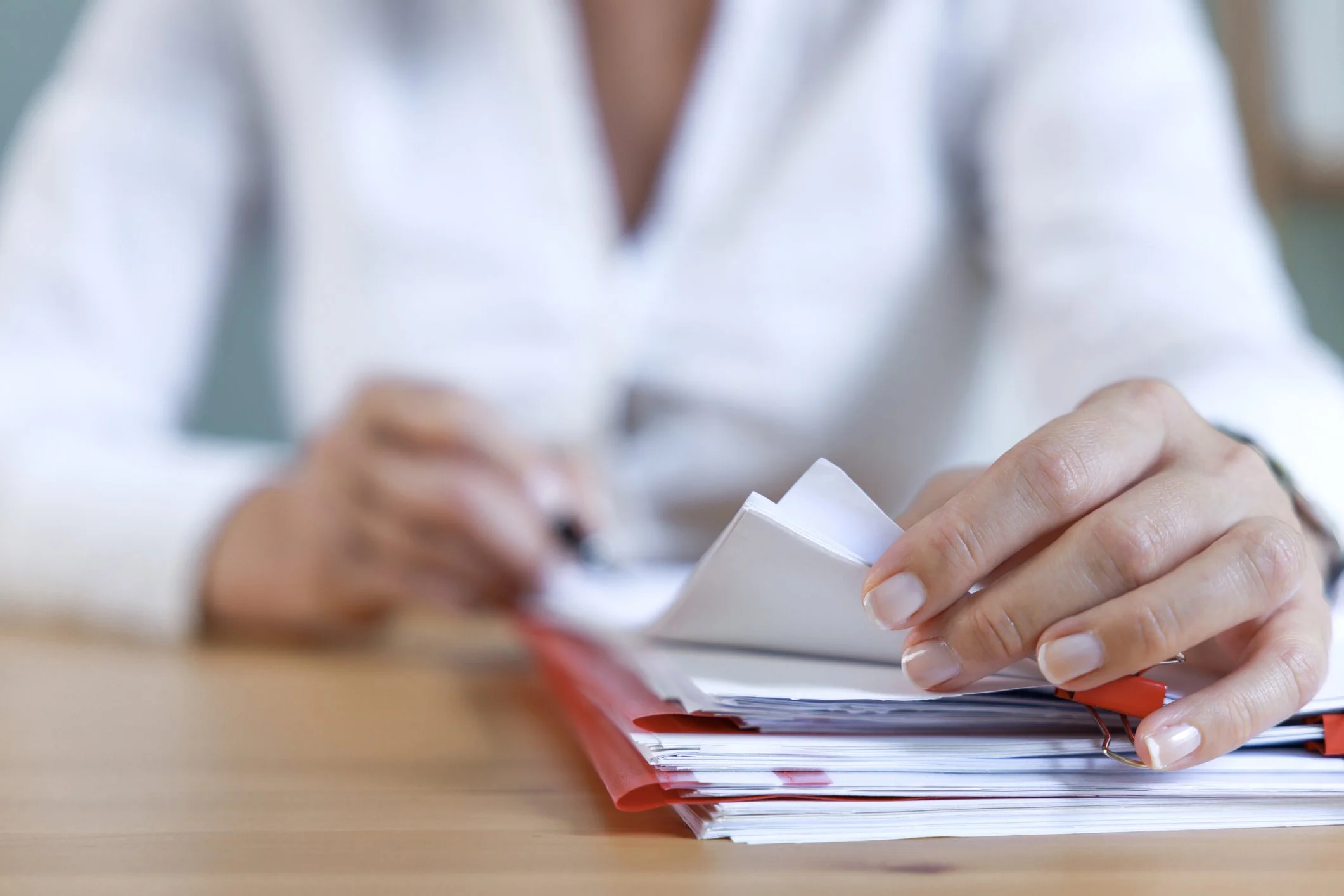 Person's hands flipping through a stack of papers or documents, with a red pen resting on top of the papers on a wooden table.