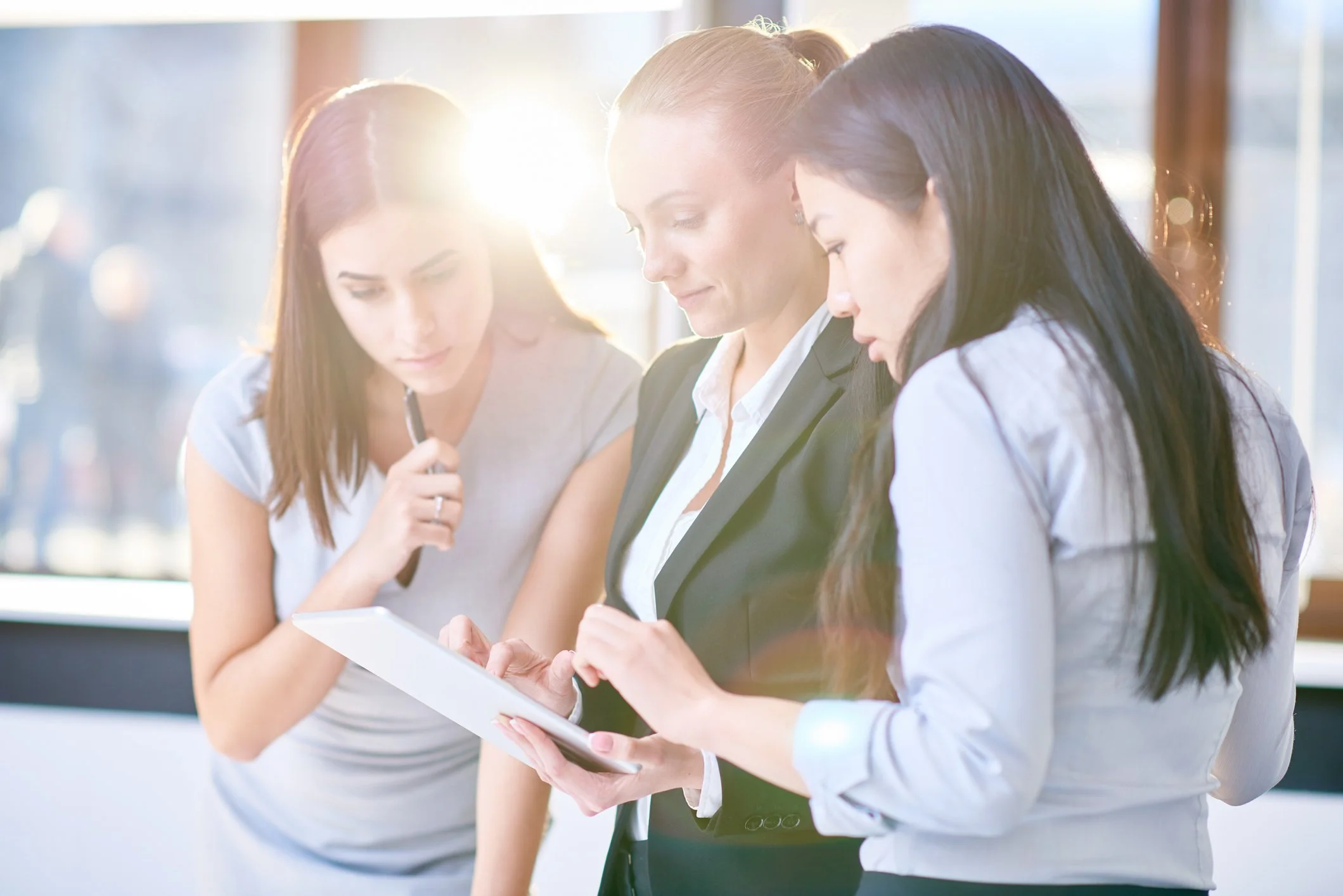 Three women are examining a document together near a window with sunlight coming through.