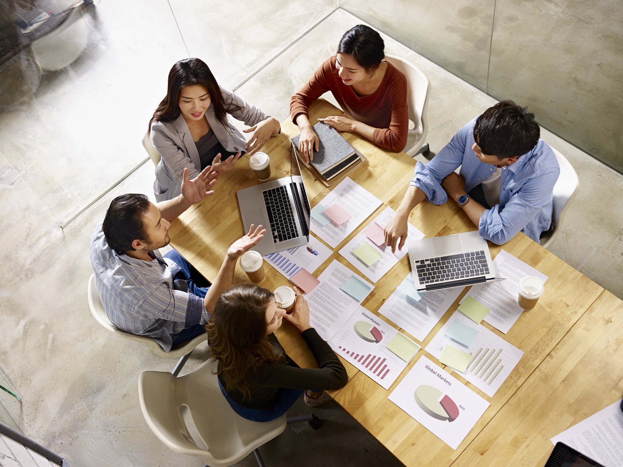A group of six diverse business professionals sitting around a wooden conference table in a meeting room. They are engaged in a discussion, with papers, charts, and notebooks spread out on the table. Some have coffee cups, and there is a silver laptop open in the center of the table. The room has a glass wall with a concrete floor.