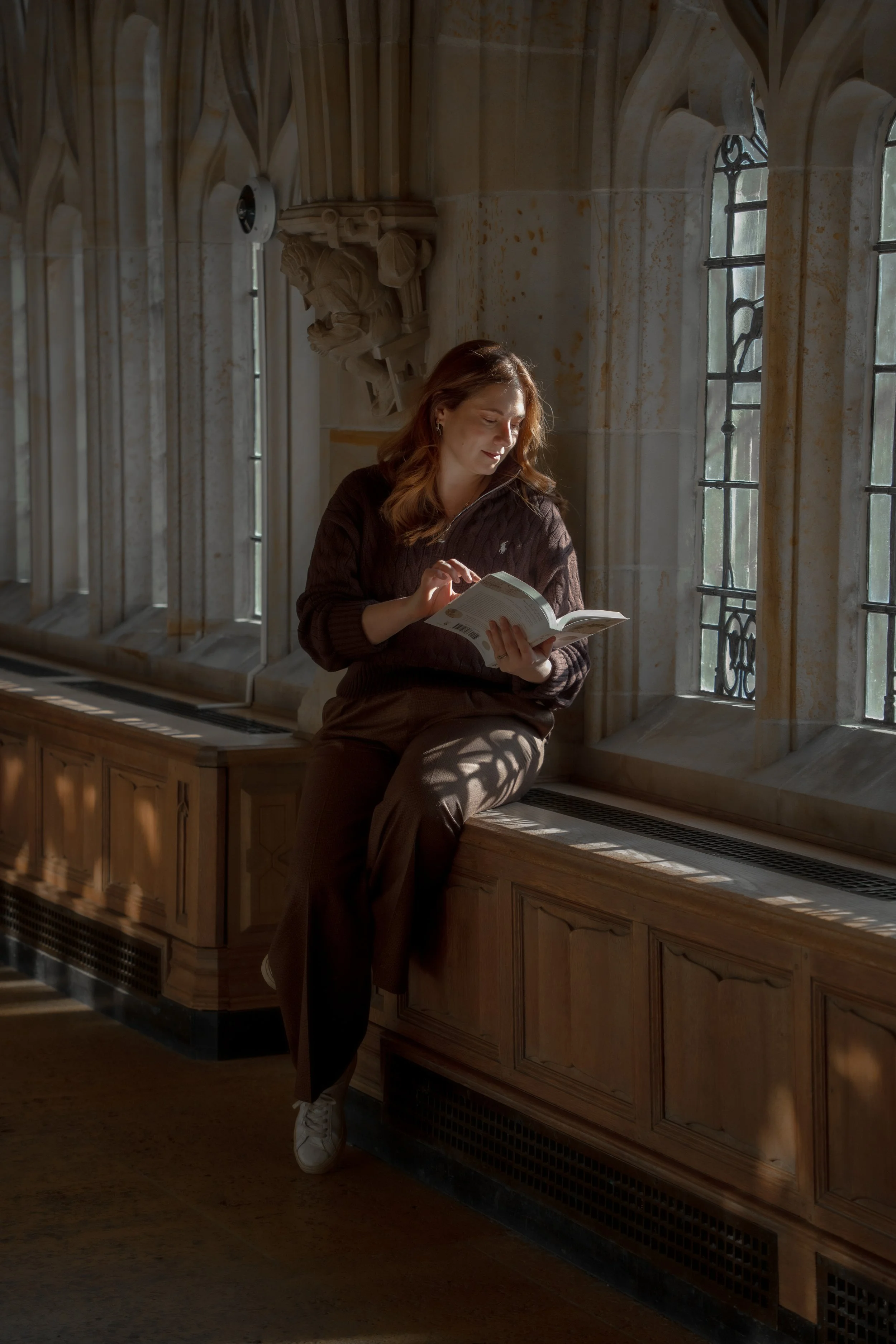 A woman with red hair reading a book, sitting on a wooden window seat in a historic stone building with tall arched windows.