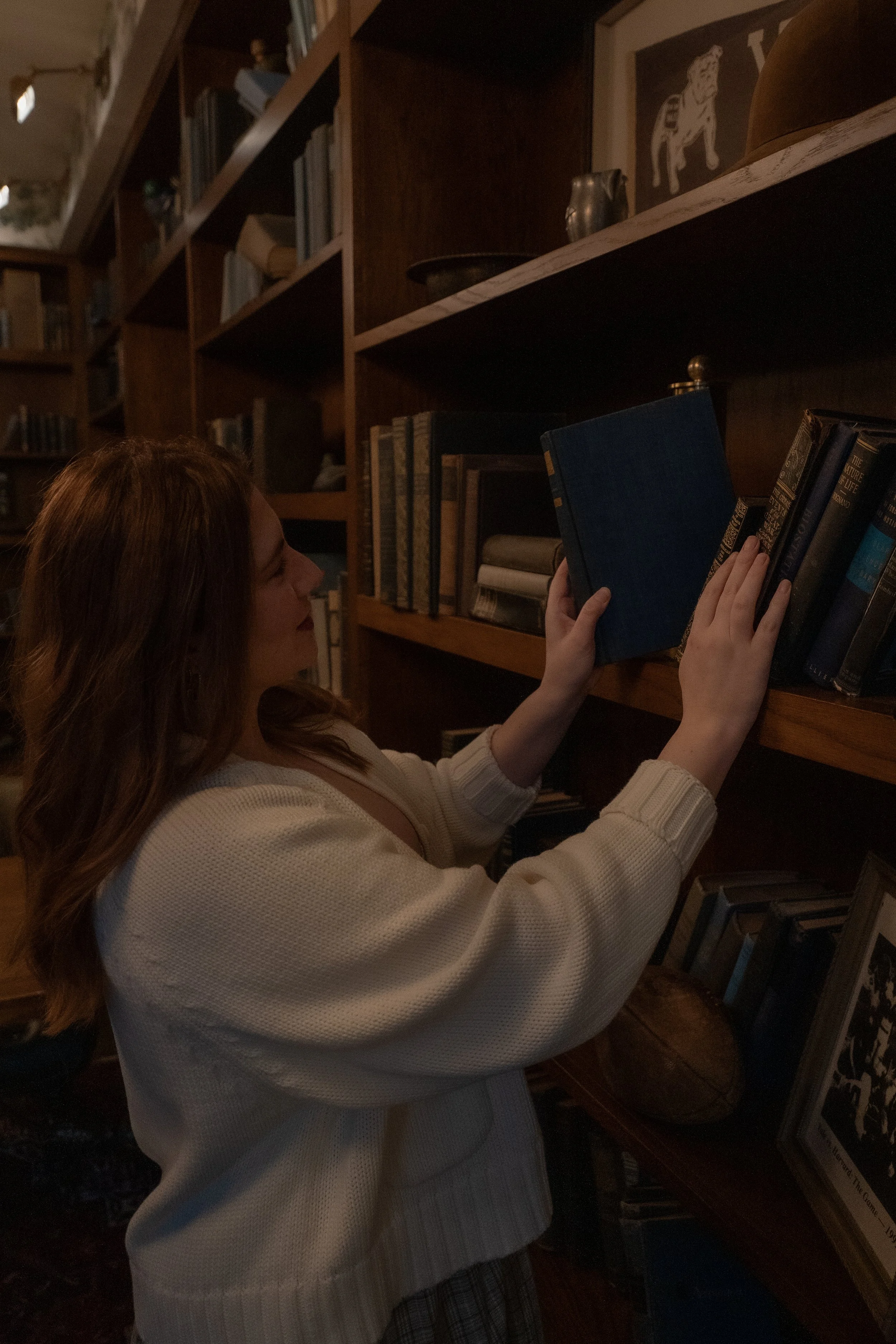 A woman with long, wavy, reddish-brown hair wearing a white sweater is browsing bookshelves in a dimly lit room, holding a blue book with her left hand while reaching for another on the shelf with her right hand.