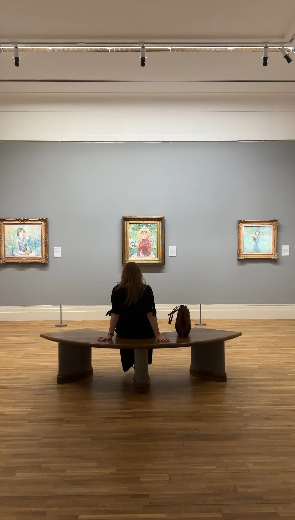 A woman sitting on a wooden bench in an art gallery, looking at three framed paintings of children on a gray wall.