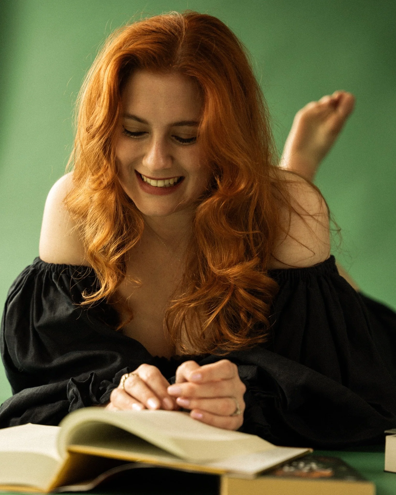 A woman with long red hair smiling while reading a book, with her hands clasped together on the table, against a green background.