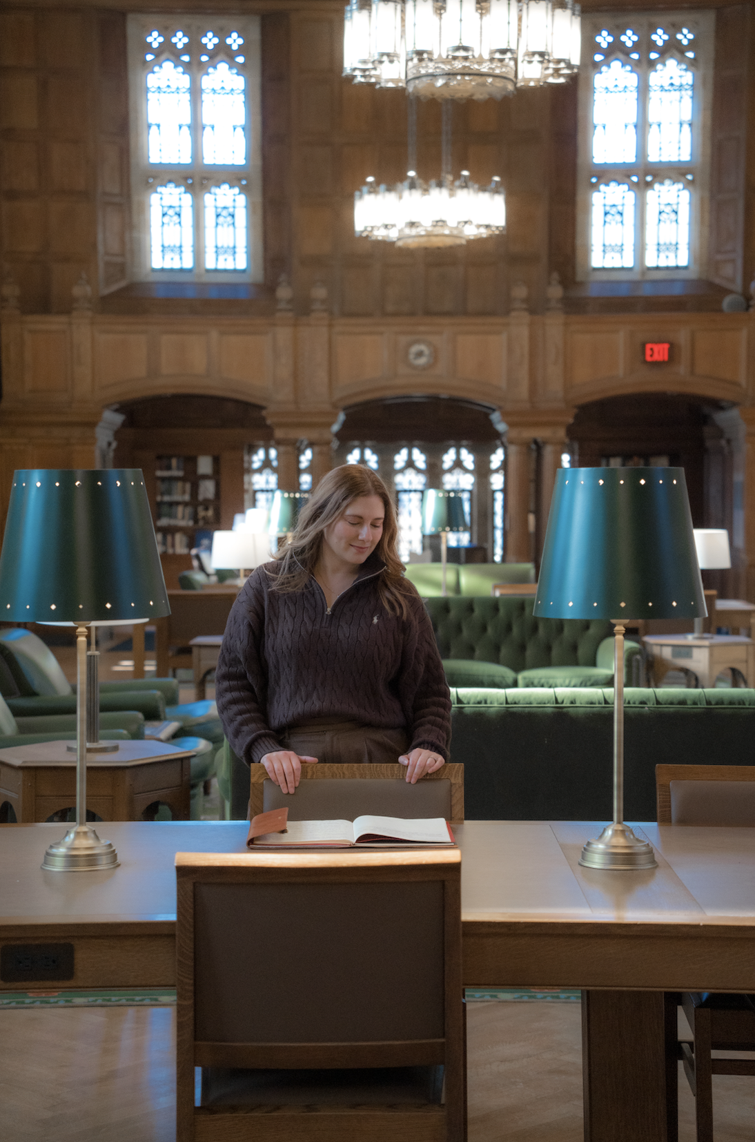 A young woman with long hair, wearing a dark sweater, stands at a wooden table in a library, looking at an open book. The library has large stained glass windows, green sofas, wooden paneling, and hanging chandeliers.