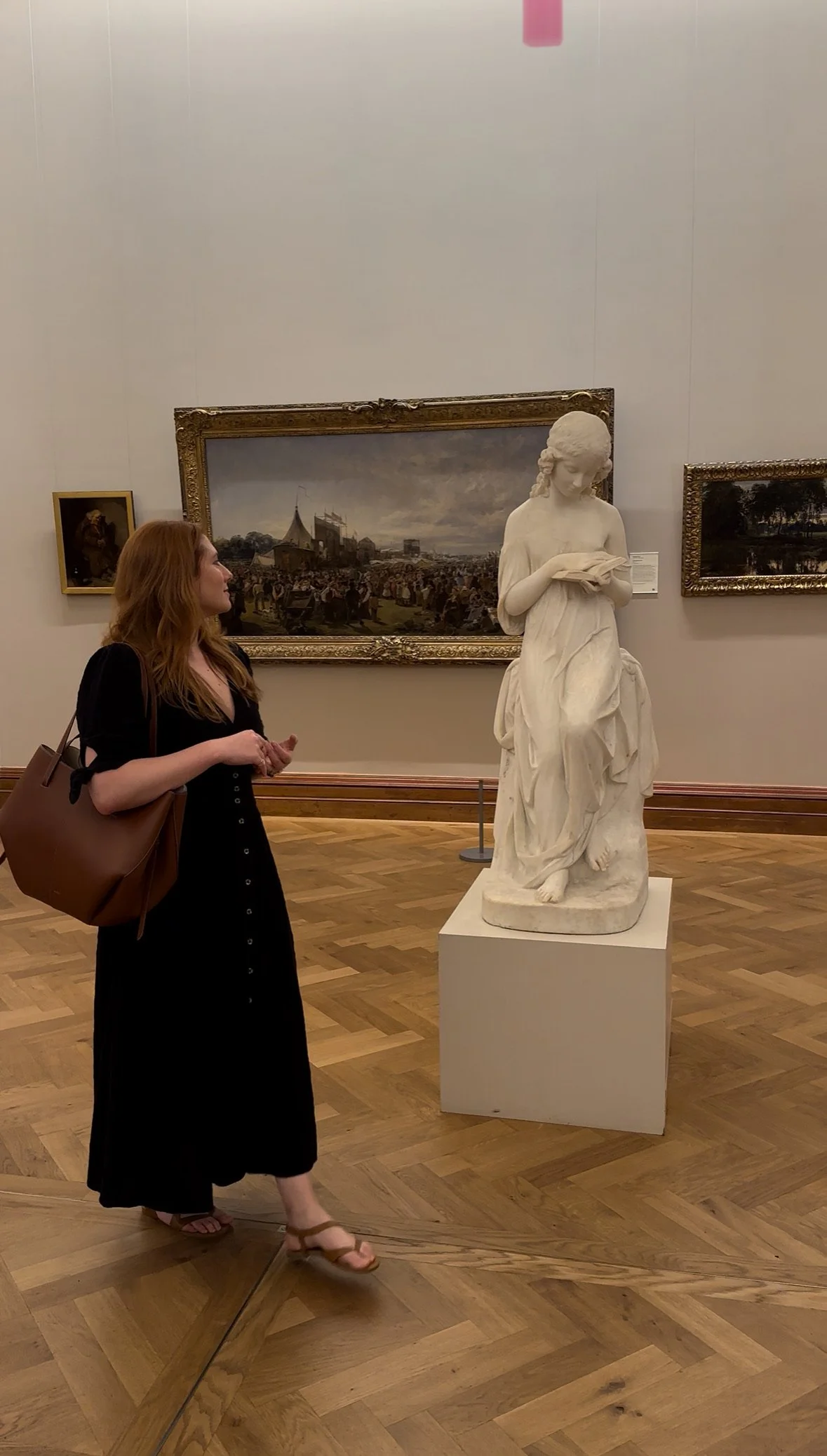 A woman with red hair wearing a black dress and brown sandals looking at a classical marble statue of a young girl reading a book in an art gallery.