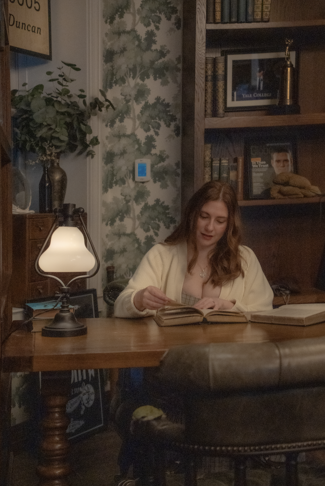 A woman with long brown hair sitting at a wooden desk in a cozy, vintage study room, reading a book with other books on the desk, illuminated by a table lamp.