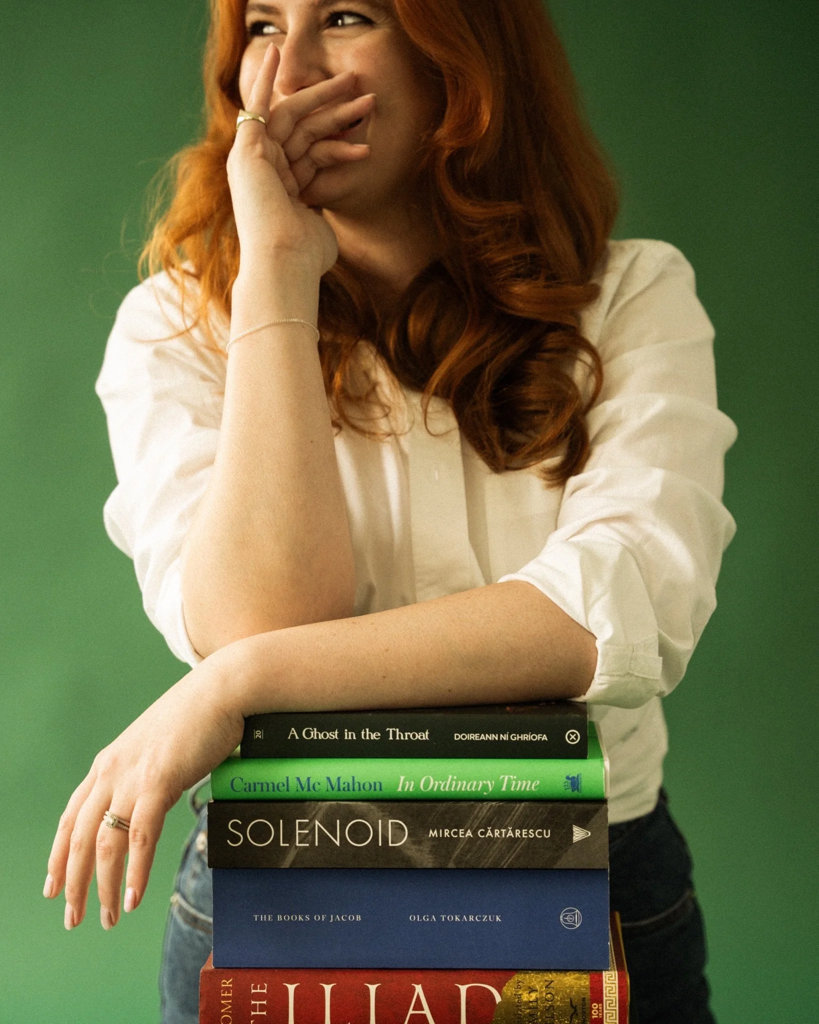 Woman with red hair leaning on a stack of books, with her hand covering her mouth and part of her face visible. The books include titles such as 'A Ghost in the Throat' by Doireann Ní Ghríofa, 'In Ordinary Time' by Carmel McMahon, 'Solenoid' by Mircea Cărtărescu, and 'The Iliad' by Homer. The background is green.
