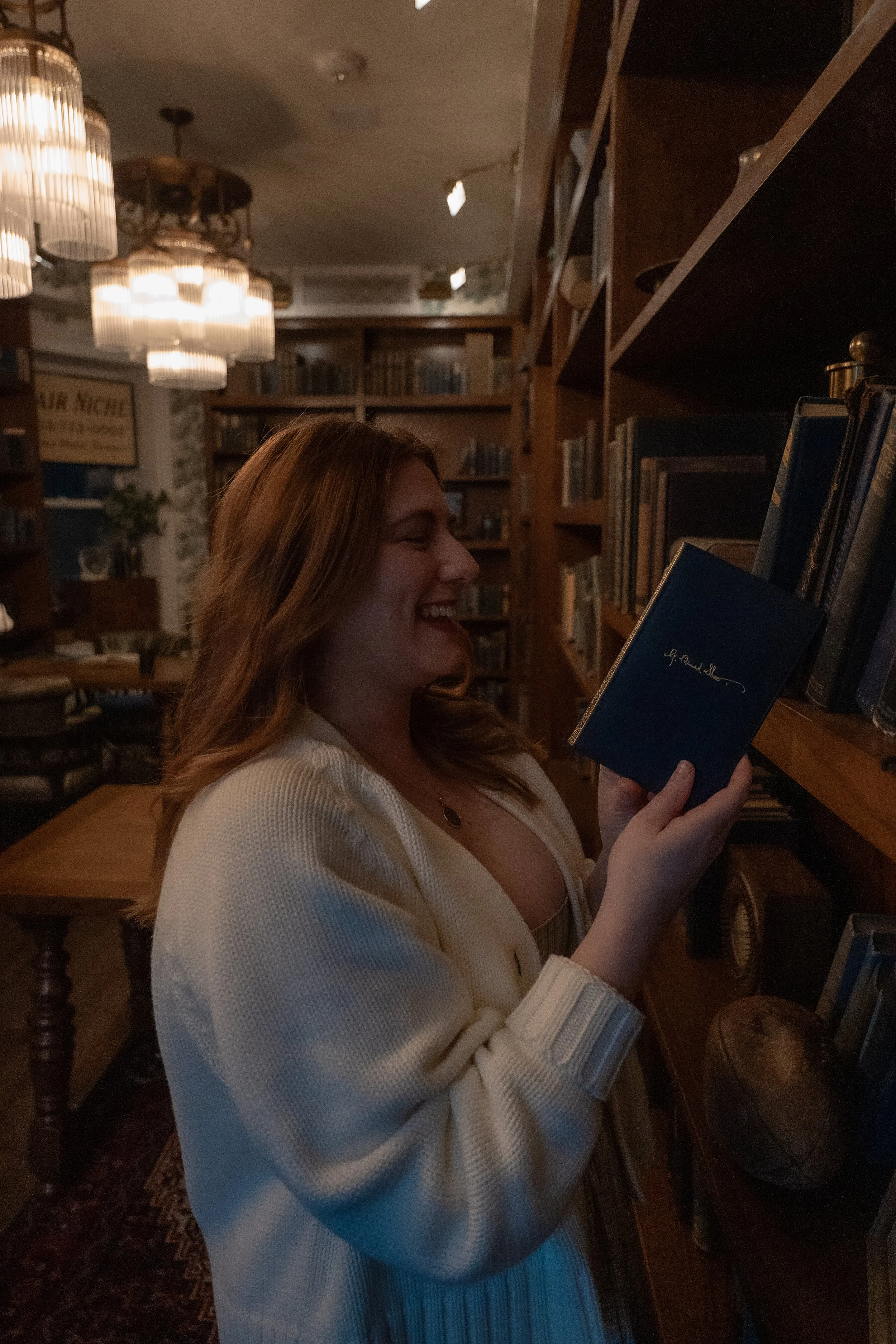 A woman with long red hair, smiling and reading a blue book in a cozy library or bookstore with wooden bookshelves and warm lighting.