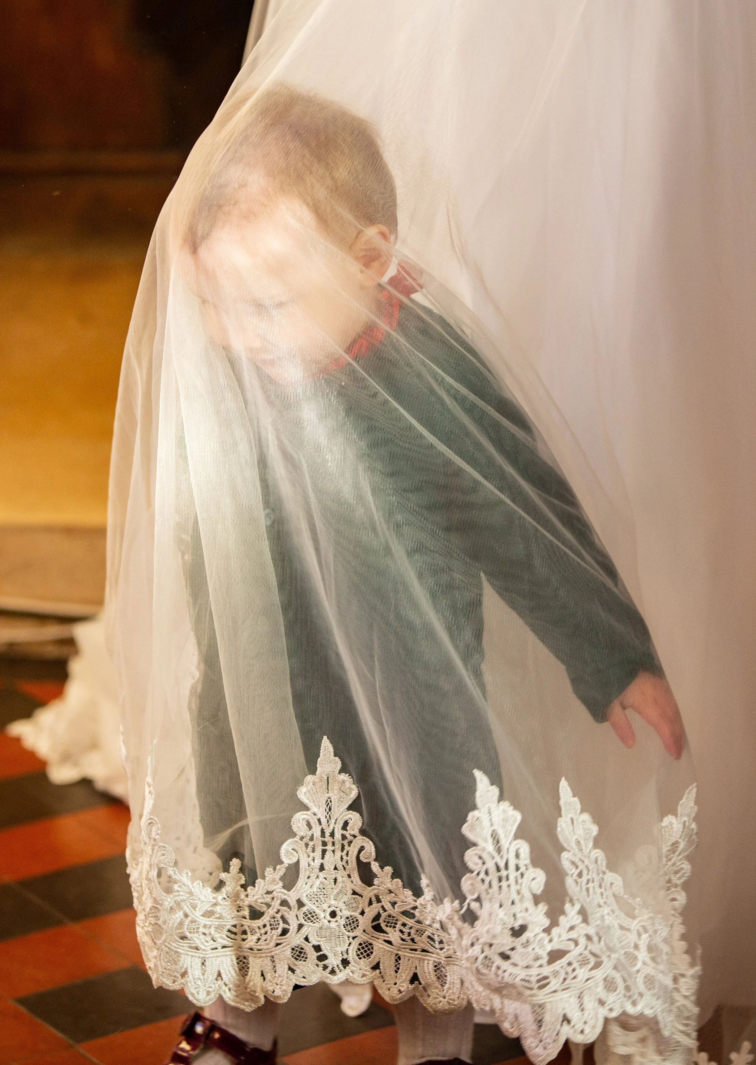 Close-up of a person wearing a lace veil with floral embroidery, standing indoors on a wooden floor.