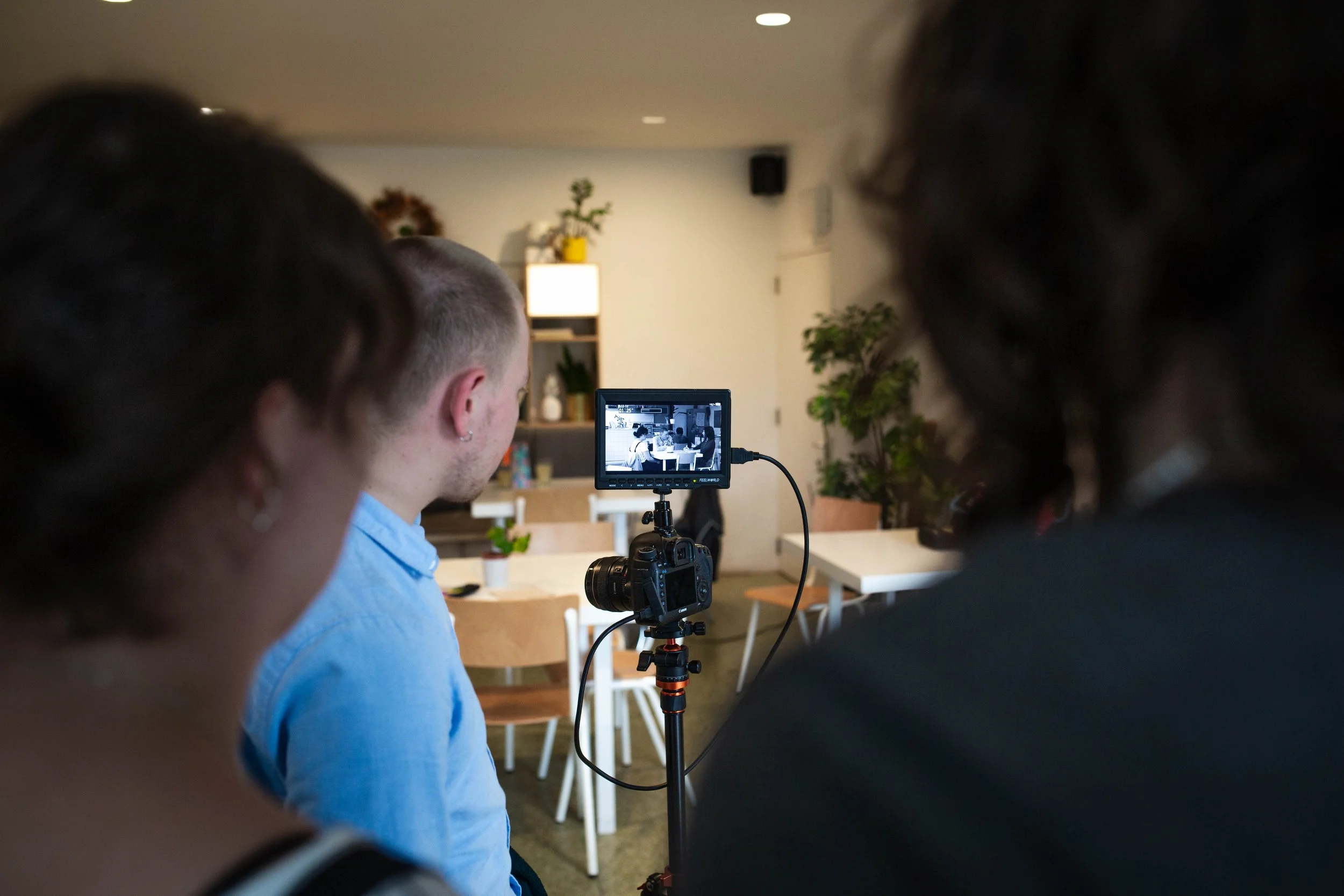 People gathered in a room watching a video being recorded by a camera on a tripod.