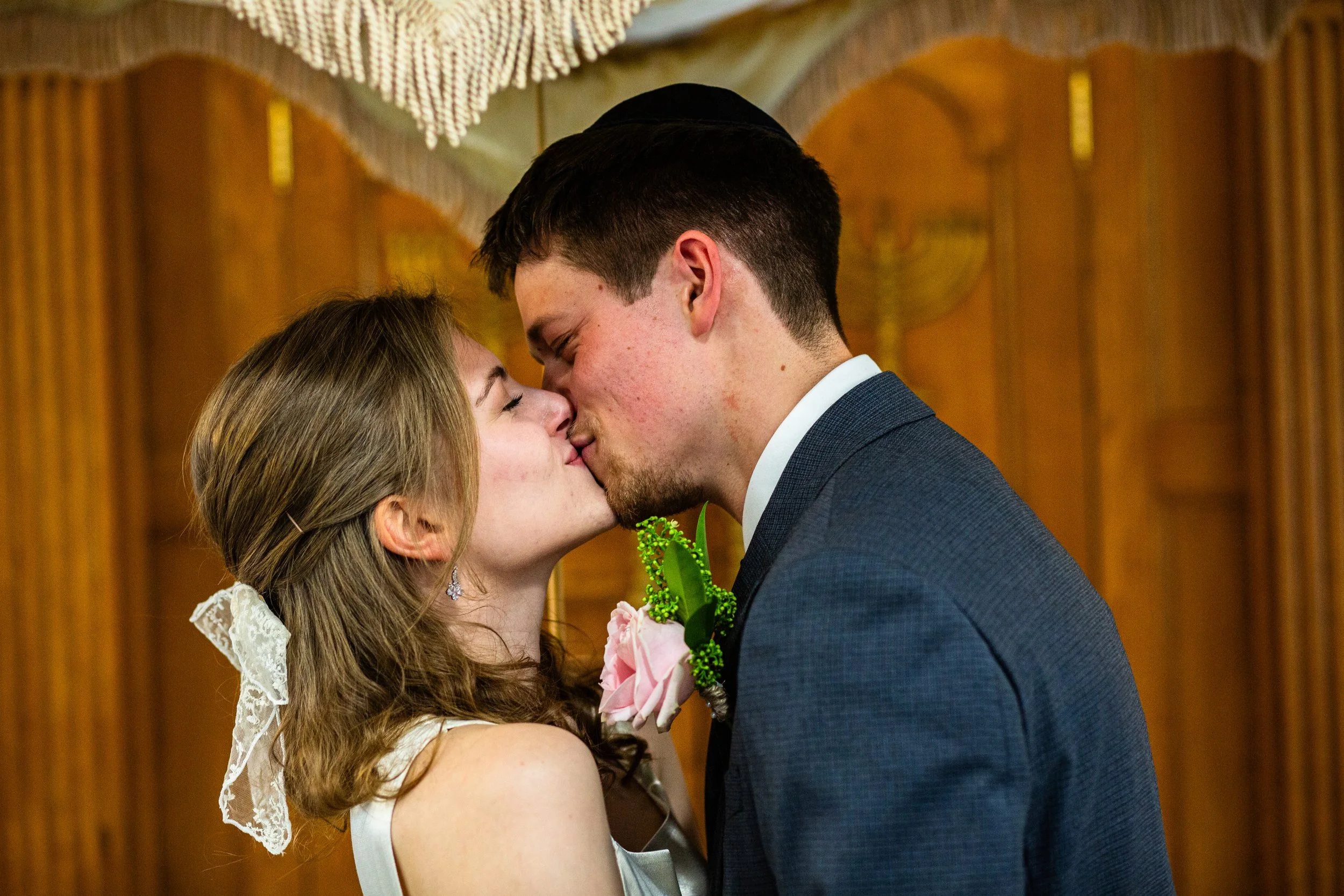A newlywed couple shares a kiss during their wedding ceremony, with the bride wearing a lace veil and the groom in a dark suit. The background features wooden panels and a decorative ceiling.