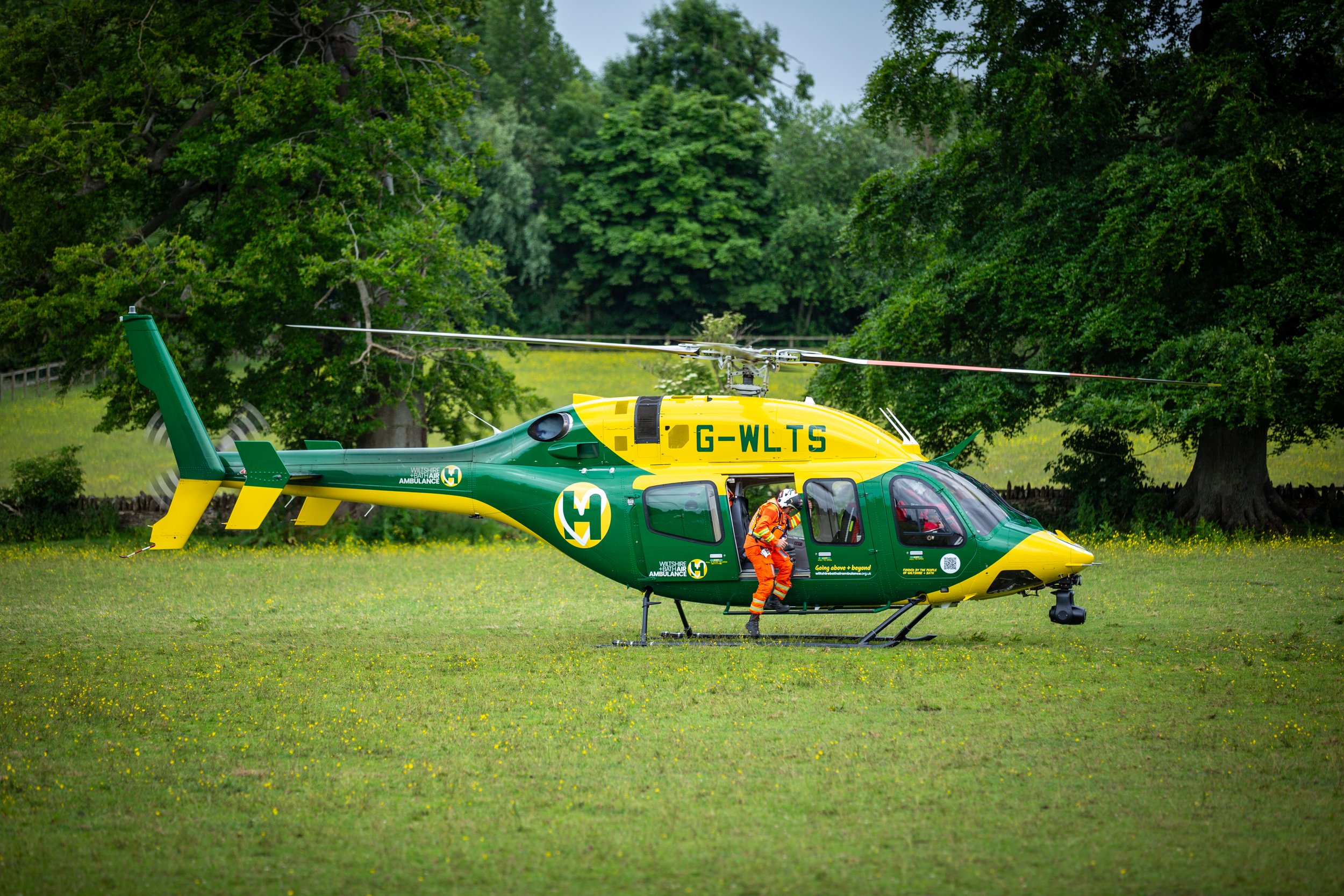 A yellow and green ambulance helicopter on a grassy field with emergency personnel. The helicopter is marked with 'Wiltshire & Bathampton Ambulance' and the registration G-WLTS, surrounded by green trees.