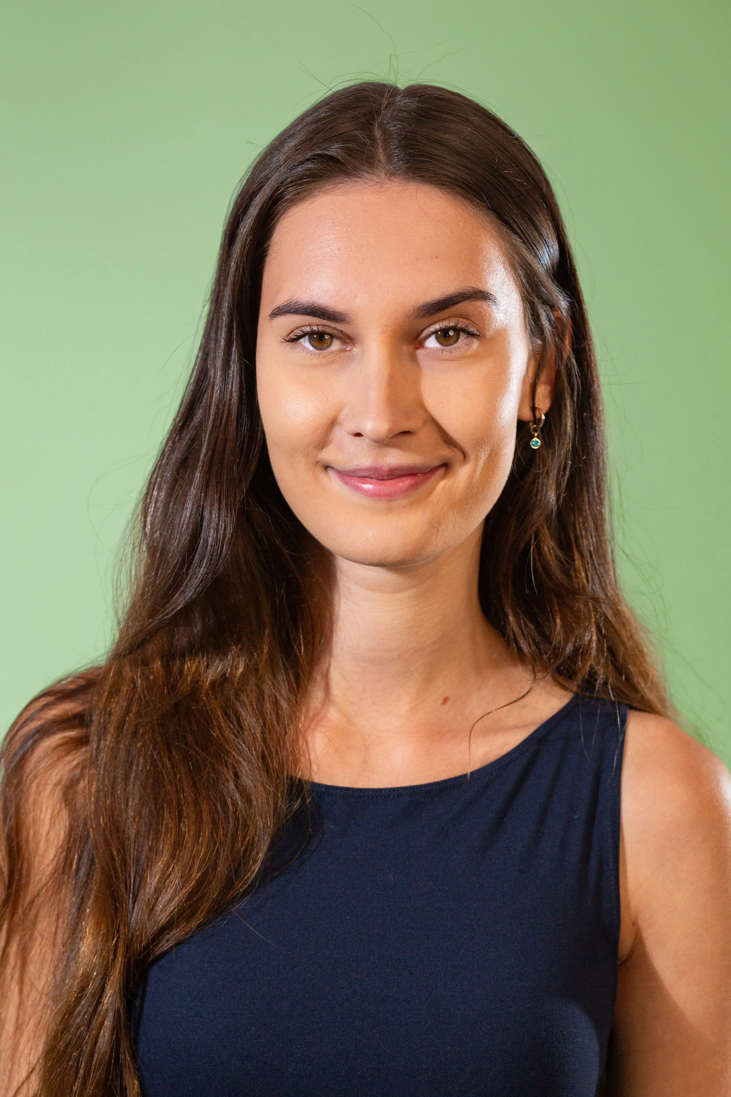 Close-up of a woman with long brown hair, hazel eyes, smiling, wearing a black sleeveless top and small earrings, against a green background. Gloucestershire.