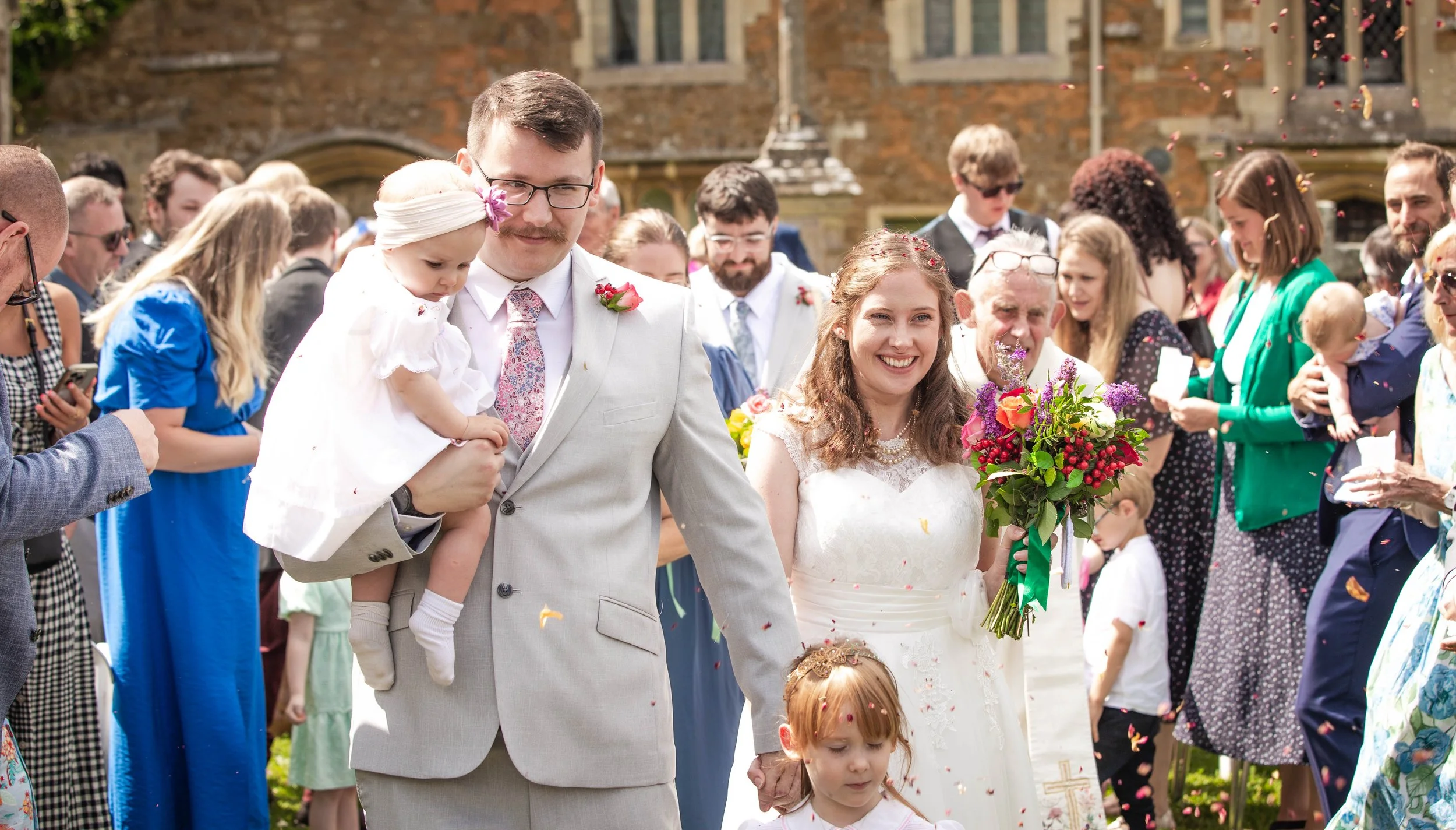 A wedding celebration with the bride and groom walking in the center, surrounded by guests, outdoors on a sunny day with falling flower petals.