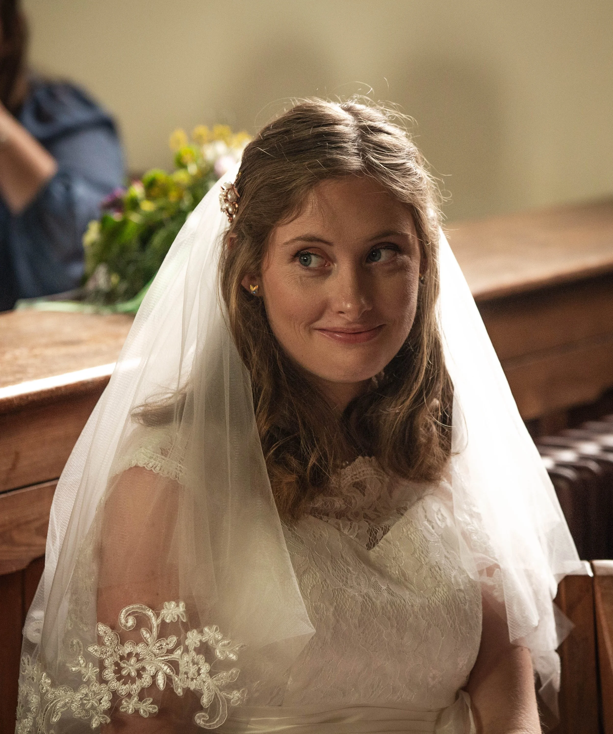 A woman wearing a white lace dress and veil, with a pearl and floral hairpiece, sitting in a church setting.