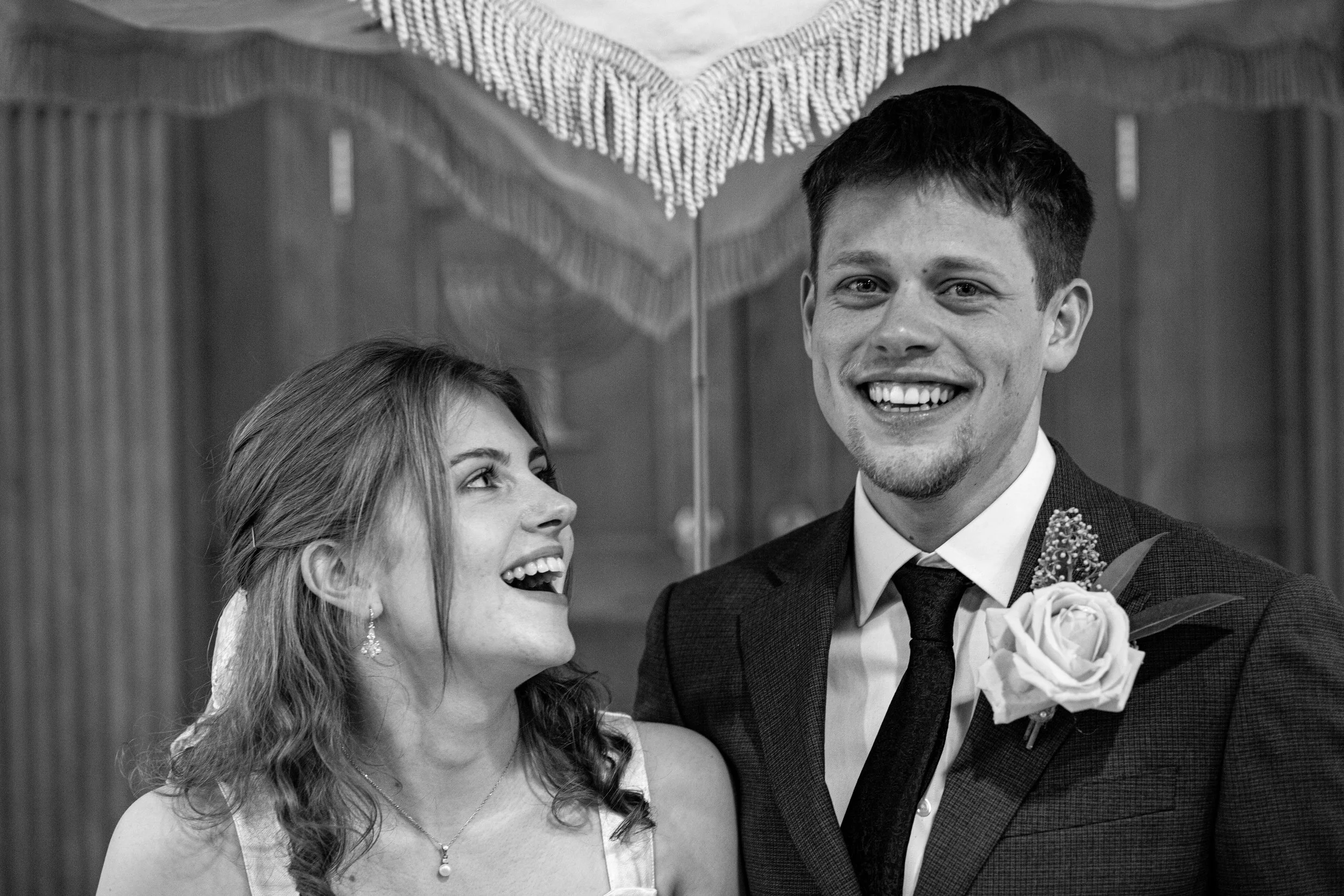 Black and white photo of a smiling man and woman at a wedding or formal event, with the woman looking up at the man. The man is wearing a suit with a boutonniere. The woman has long wavy hair and is wearing earrings and a necklace.