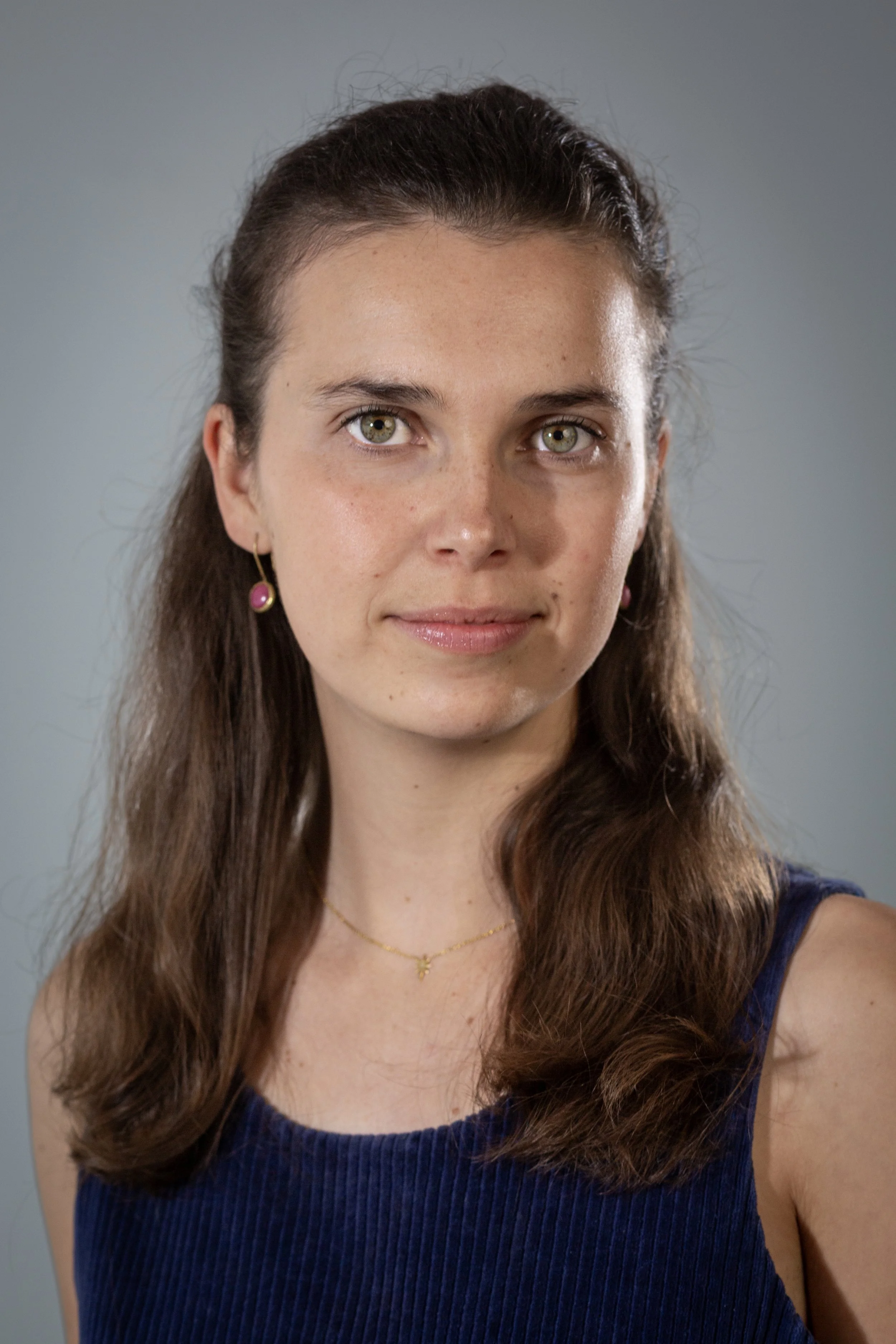 Close-up portrait of a young woman with long brown hair, light green eyes, wearing pink earrings, a gold necklace with a small pendant, and a dark blue sleeveless top, against a plain grey background. Gloucestershire.