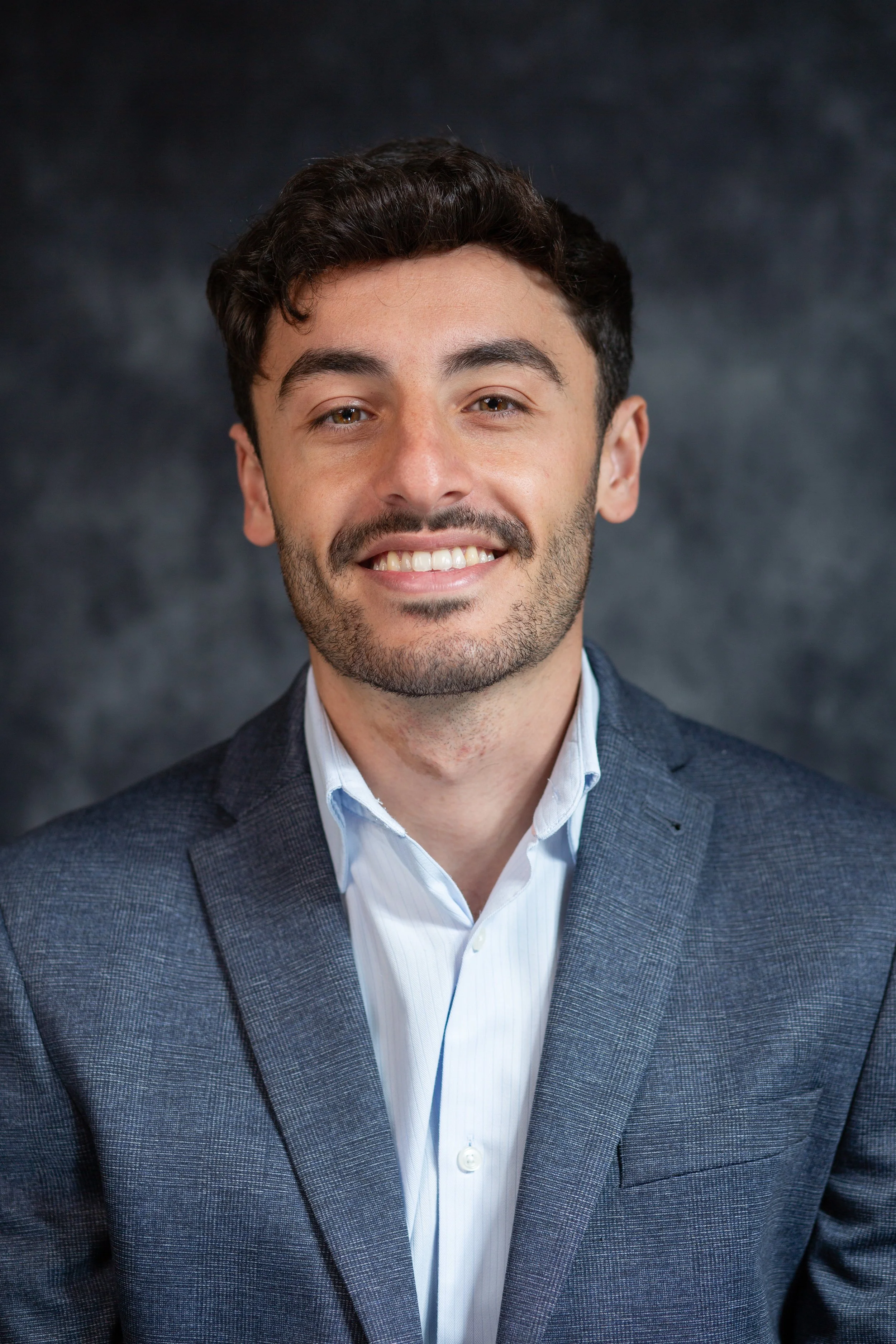 Portrait of a young man with dark hair, brown eyes, light skin, wearing a dark blue checked blazer and a white shirt, smiling against a dark textured background. Gloucestershire.
