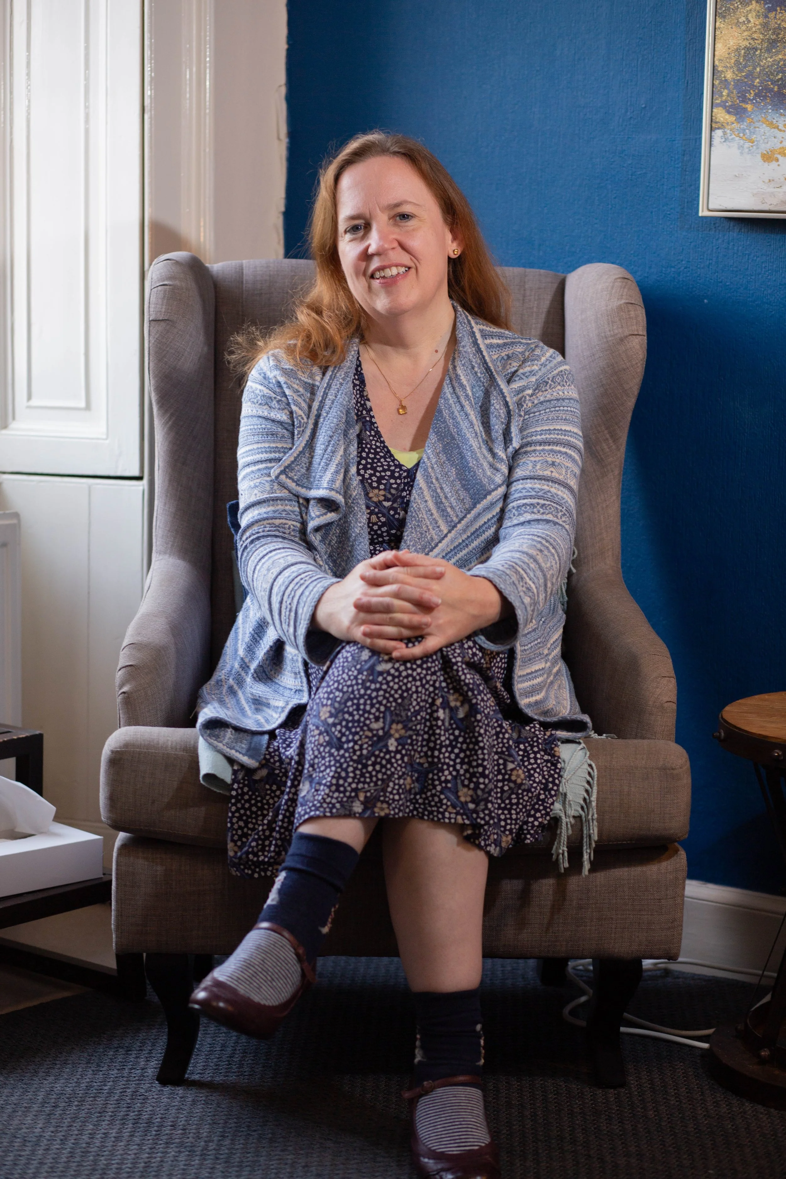 A woman with red hair sitting in a beige armchair in a living room, smiling at the camera, with blue and white walls and a piece of artwork on the wall behind her. Wotton Under Edge, Gloucestershire