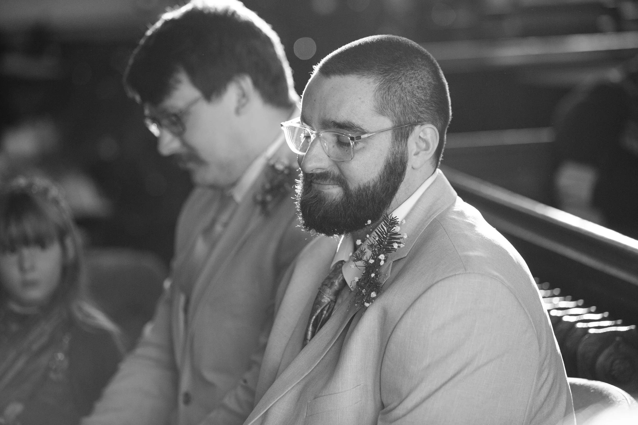 Black and white photo of two men with glasses, dressed in suits, sitting closely with heads bowed and eyes closed, possibly during a formal event or ceremony.