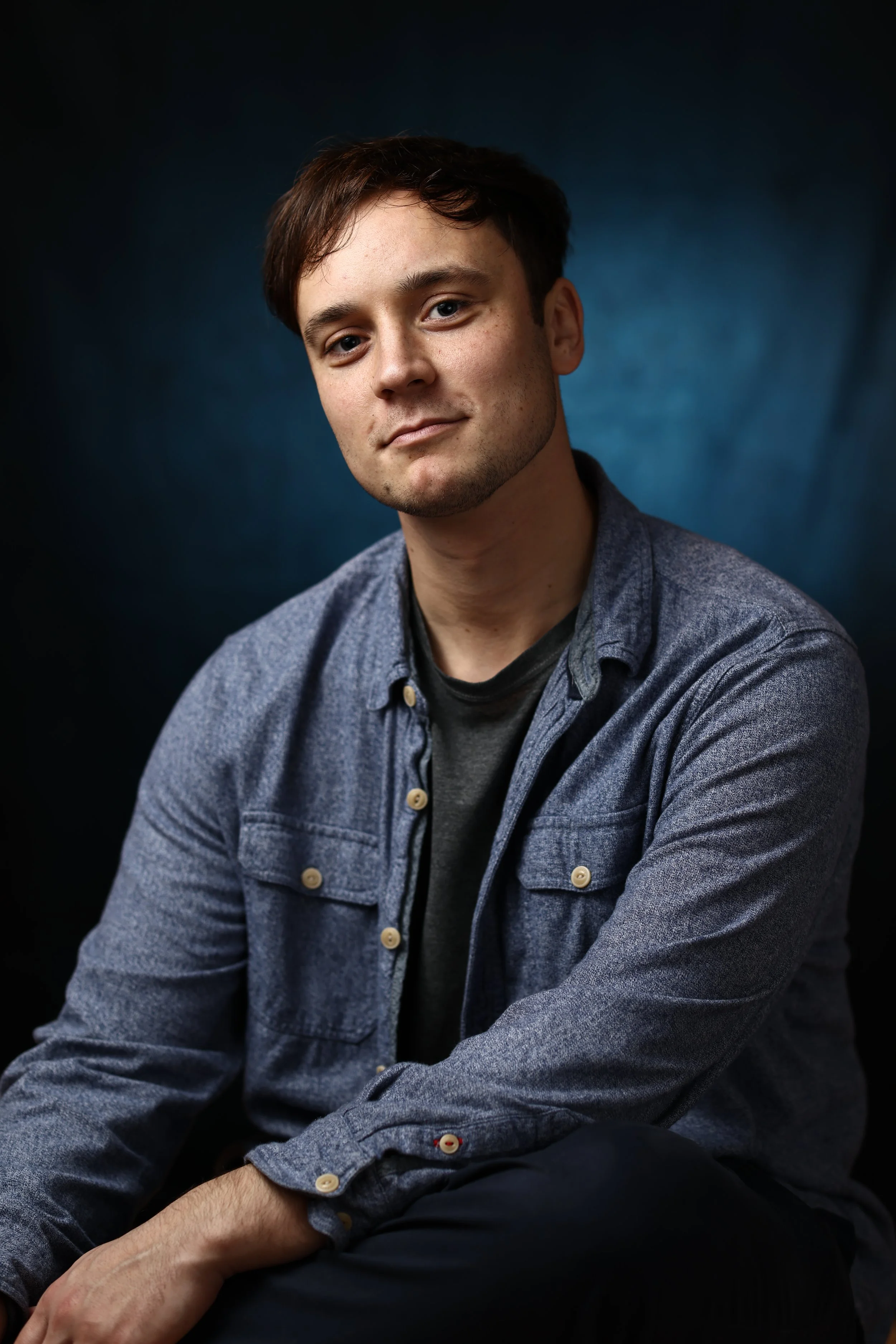 Portrait of a young man with short brown hair and blue eyes, wearing a denim button-up shirt, sitting with his arms crossed, against a dark blue background.