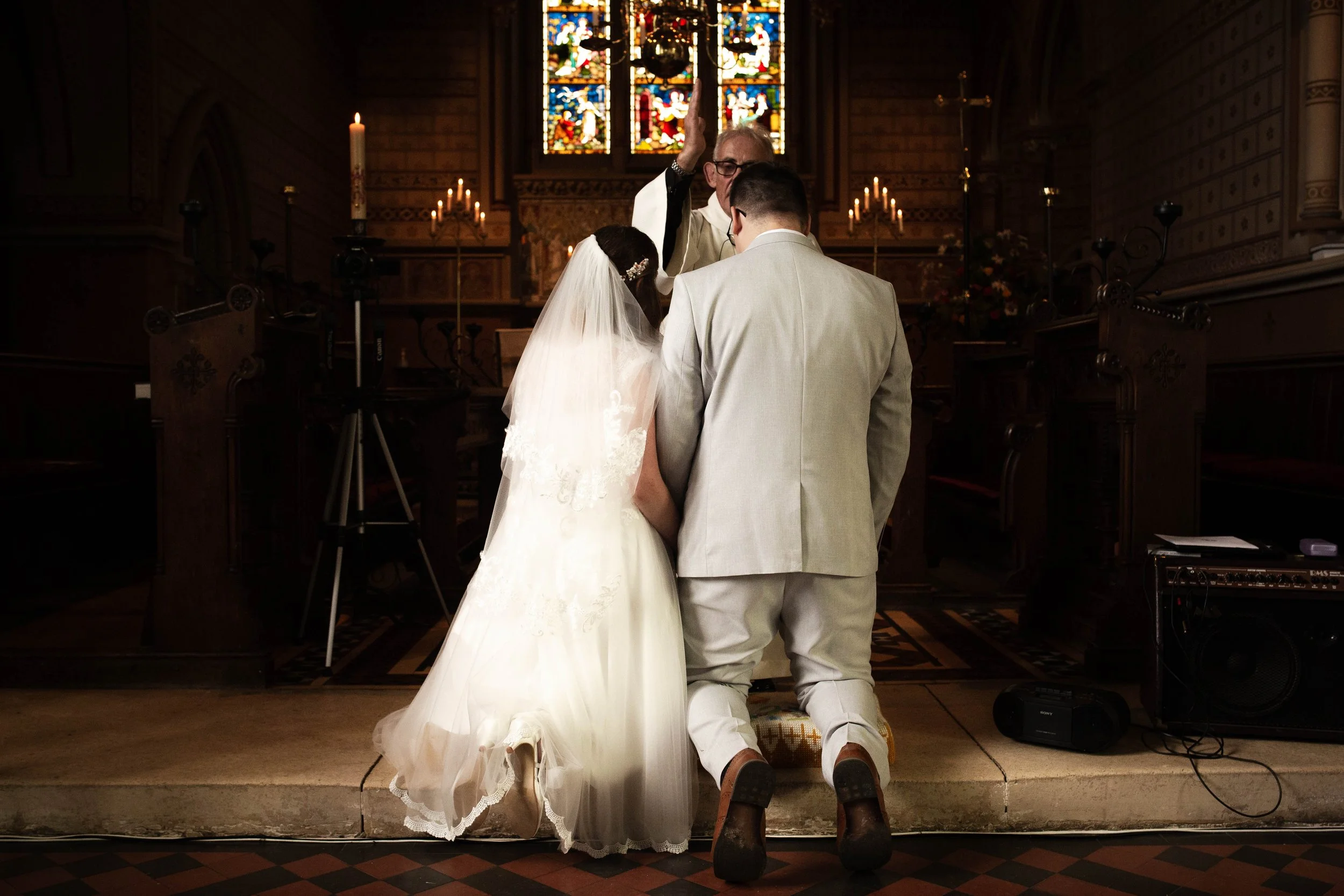 A wedding ceremony inside a church with three people kneeling before a priest performing the vows. The bride is wearing a white wedding gown and veil, the groom in a light-colored suit, and the officiant in religious robes. The church has stained gla
